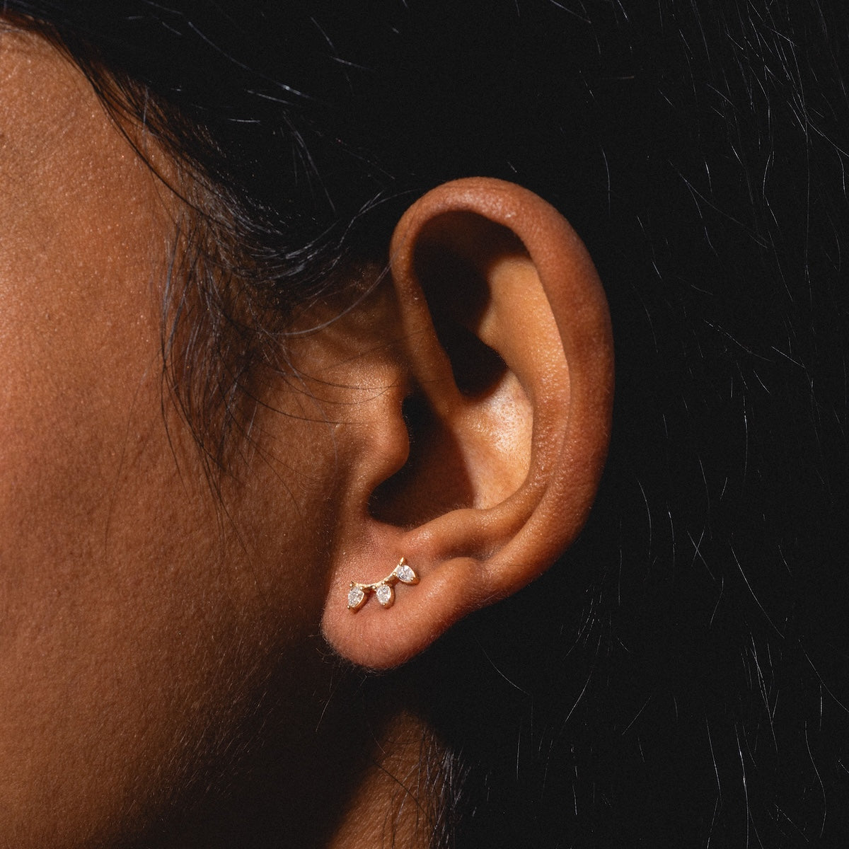Close-up of an ear adorned with three Rain Diamond Studs—small gold and diamond earrings arranged in a line along the earlobe. A dark background highlights both the elegant jewelry and the shape of the ear.