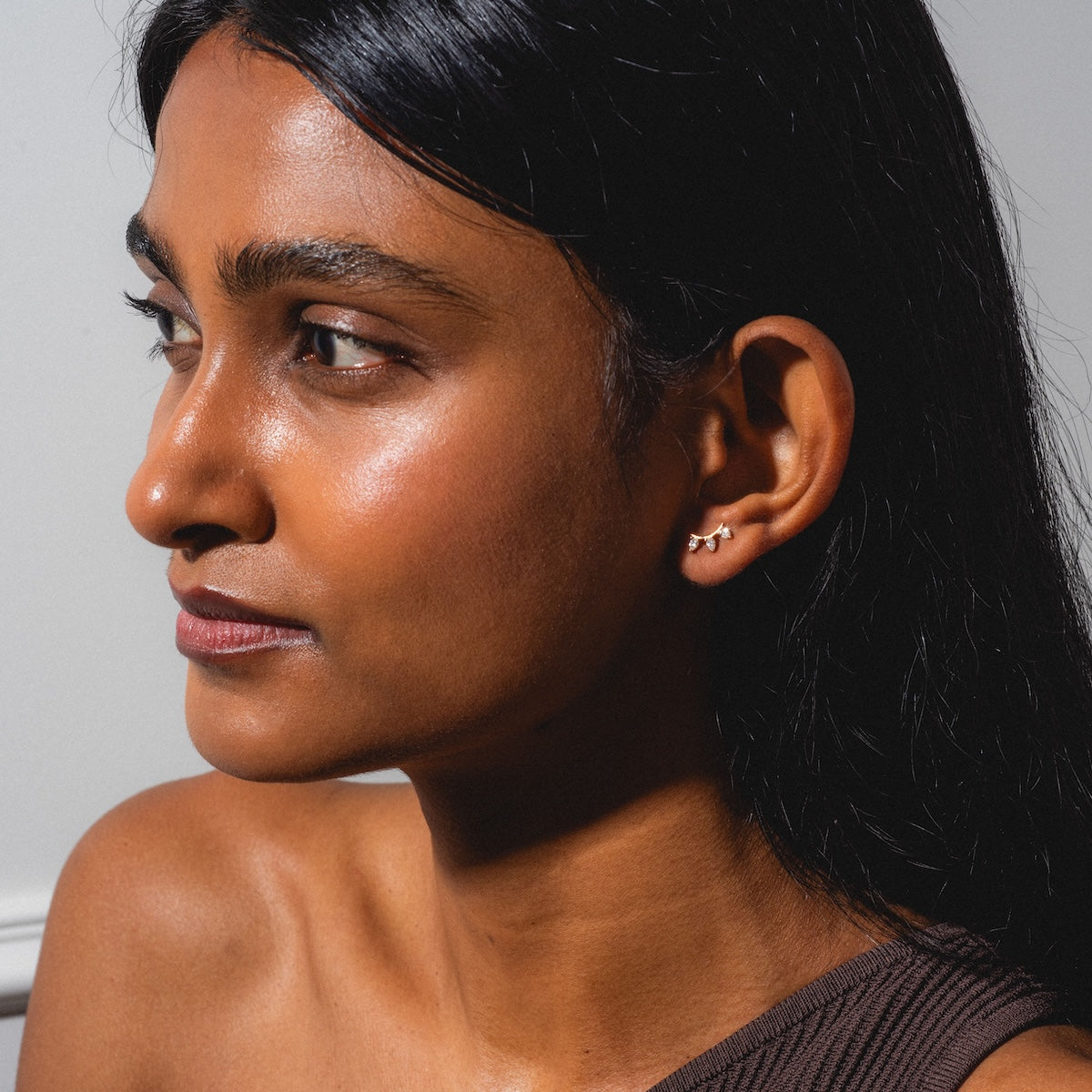 A woman with long dark hair and radiant skin poses in profile against a light background, wearing a sleeveless brown top and Rain Diamond Studs earrings.