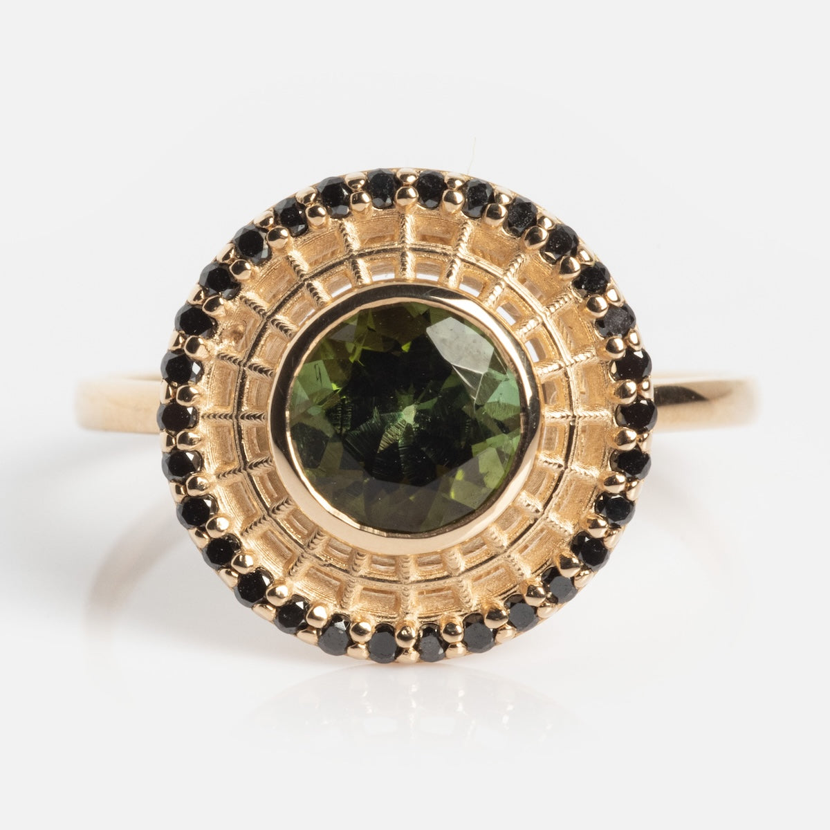 The Tantallon Black Diamond Ring features a central round green gemstone, encircled by a patterned gold halo and bordered with small black diamonds. The ring is displayed on a plain white background.
