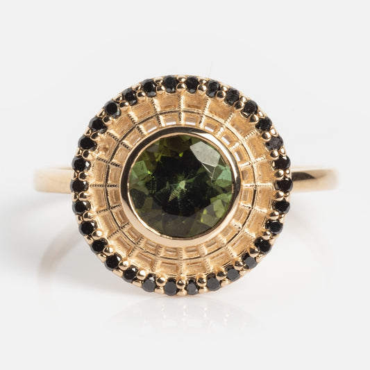 The Tantallon Black Diamond Ring features a central round green gemstone, encircled by a patterned gold halo and bordered with small black diamonds. The ring is displayed on a plain white background.