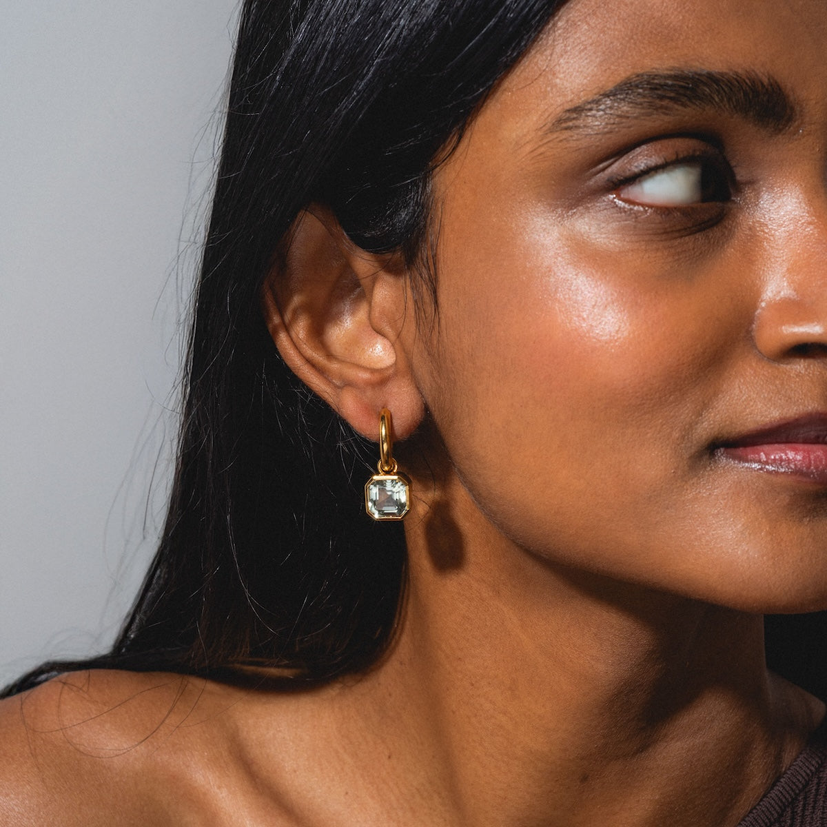 A woman with smooth brown skin and long dark hair gazes to the side, wearing Green Amethyst Drop Hoop Earrings. The plain background and soft lighting enhance her glowing complexion and the elegance of the earrings.