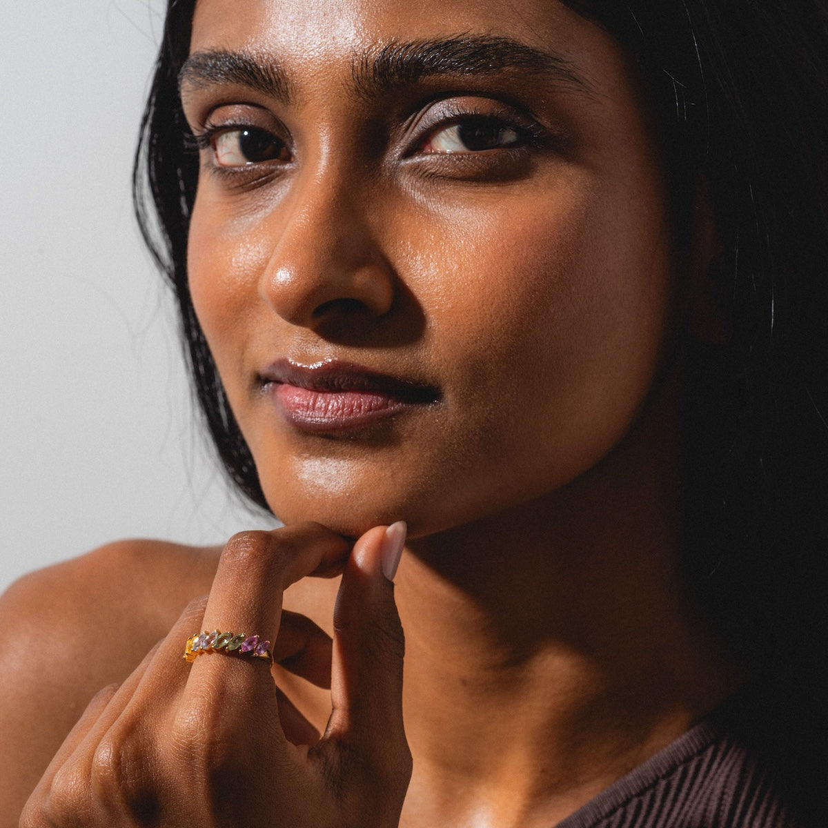 A woman with long dark hair and glowing skin gazes at the camera, gently touching her chin. She wears the Della Rainbow Sapphire Ring and a brown off-shoulder top, bathed in soft natural light.