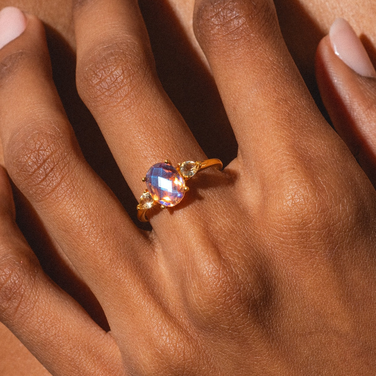 A close-up of a hand with smooth brown skin and manicured nails wearing the Pippa Quartz Ring, featuring a large iridescent gemstone flanked by smaller stones on each side.