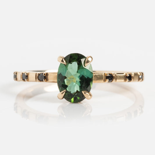 The Iona Ring is a 14k yellow gold band featuring a large oval green center gemstone, accented by small black diamonds along the thin band, shown on a plain white background.
