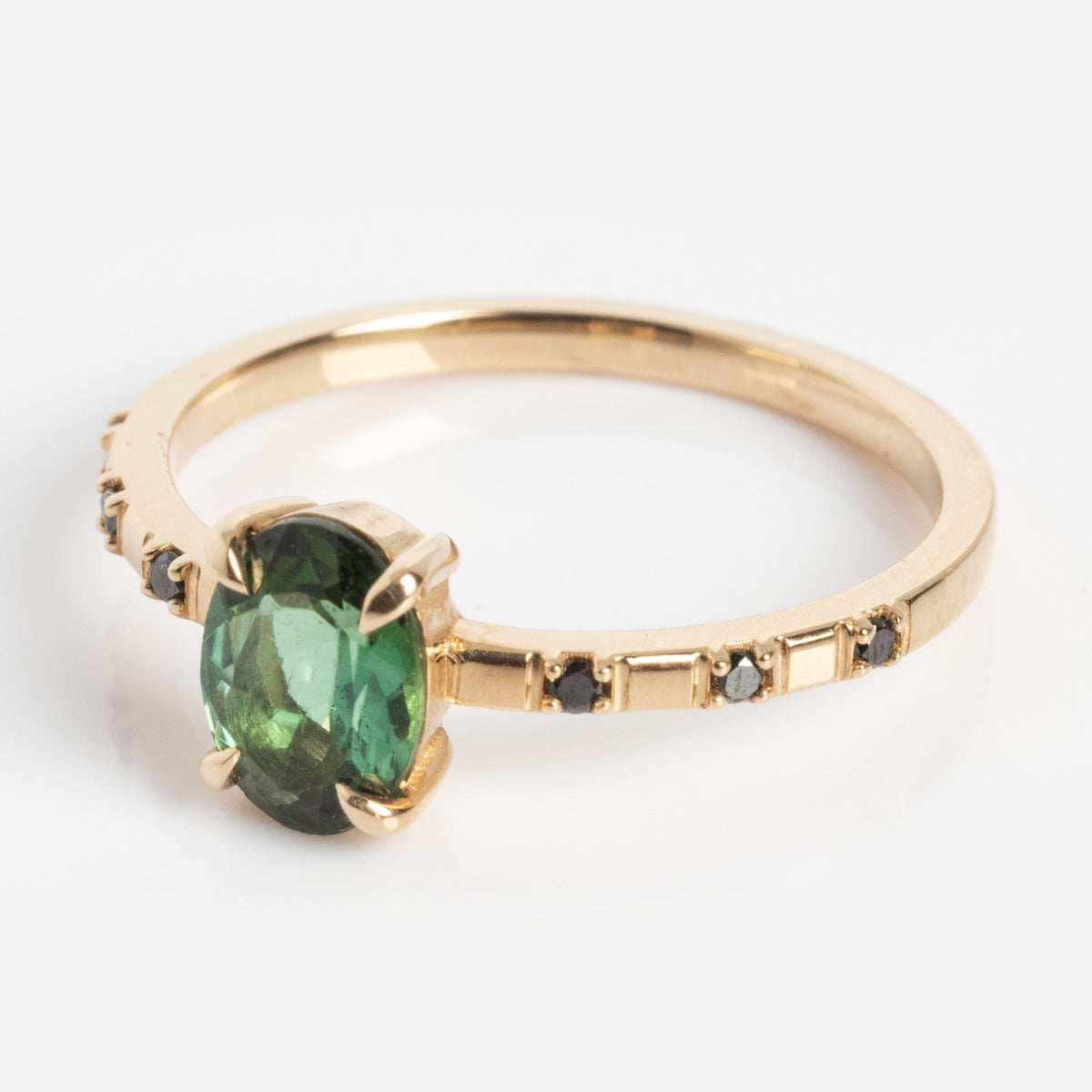 The Iona Ring Black Diamond Ring is a 14k yellow gold ring with a large round blue-green tourmaline center stone and small black diamonds set along one side of the band, shown on a plain white background.