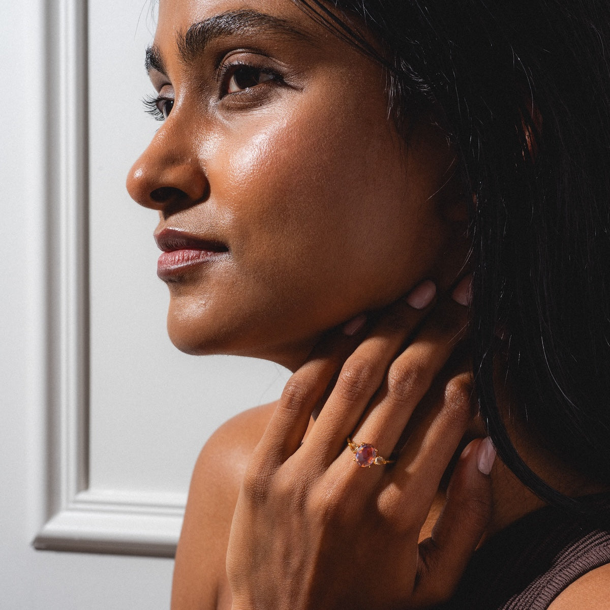 A woman with radiant skin gazes thoughtfully to the side, her hand touching her neck and chin to showcase the Pippa Quartz Ring—a gold band set with a pink gemstone—while standing before a white paneled wall.