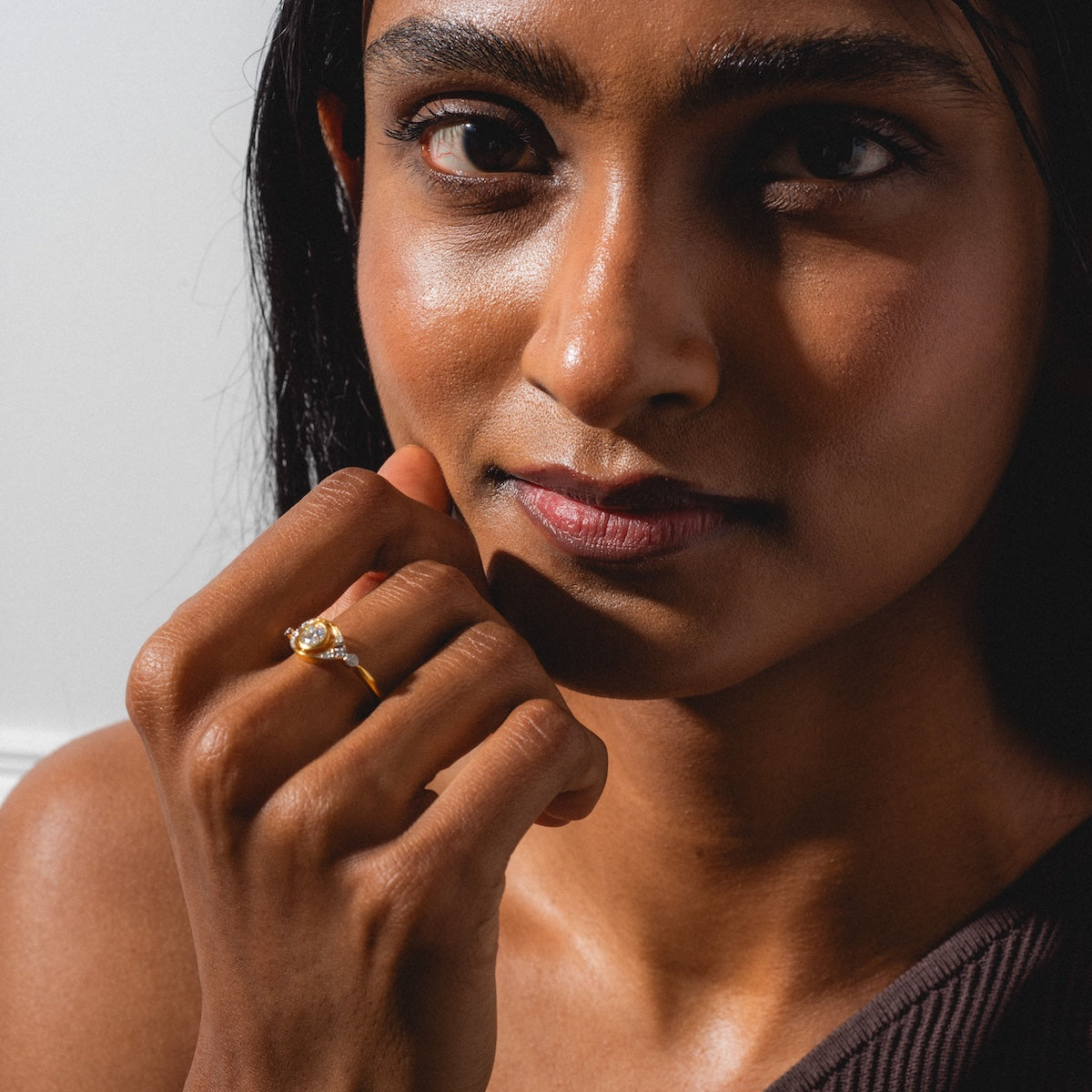 A woman with long dark hair gazes at the camera, displaying the Vienna Moissanite and Diamond Ring on her hand. She wears a brown one-shoulder top, illuminated by natural light.
