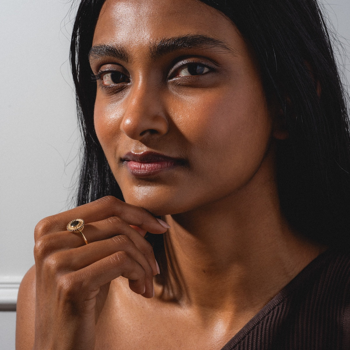A woman with long dark hair, wearing a one-shoulder dark brown top, gazes at the camera while resting her chin on her hand and showcasing the Tantallon Black Diamond Ring.