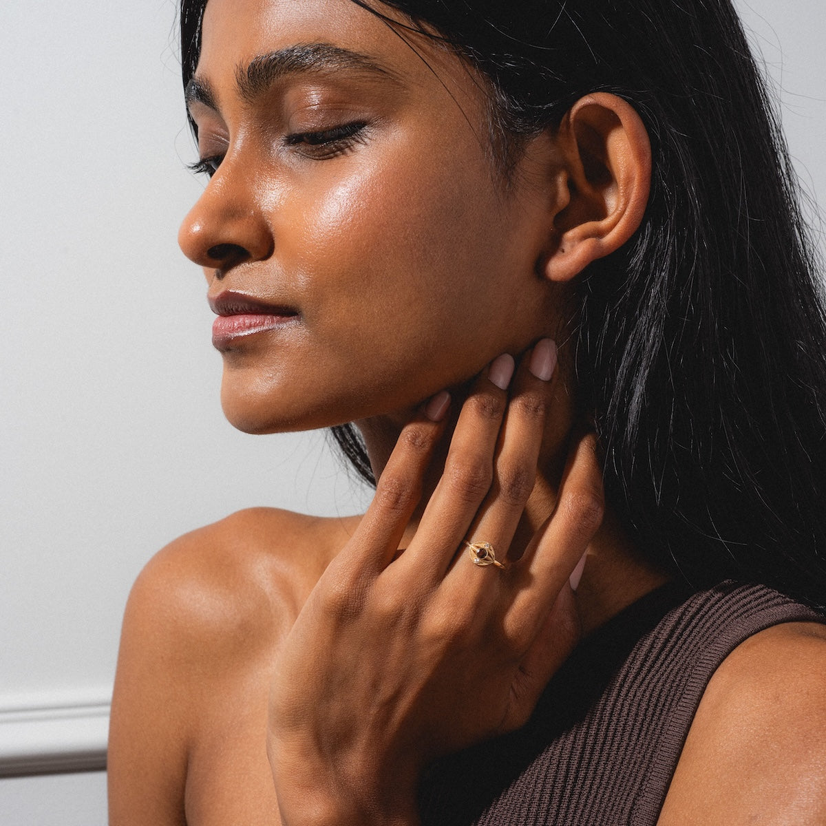 A woman with long dark hair and glowing skin closes her eyes, gently touching her neck while wearing a brown one-shoulder top and the Staffa White Diamond Ring against a plain light background.