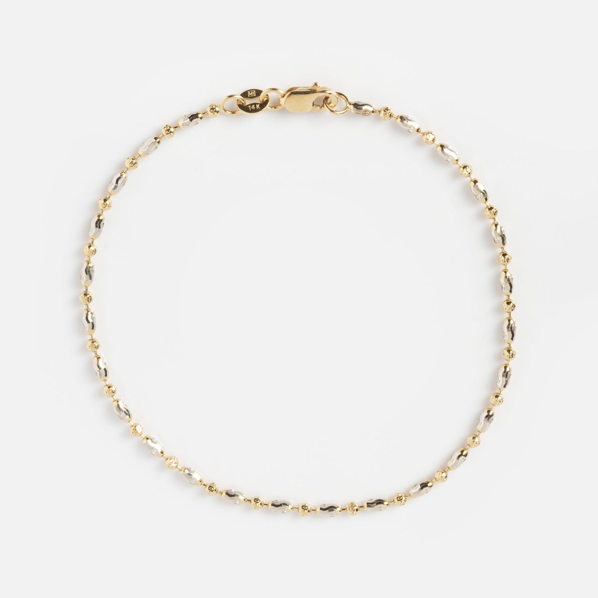 The Bubble Two Tone Bracelet is a delicate gold chain with small bead accents and a lobster clasp, displayed in a circular shape on a plain white background.