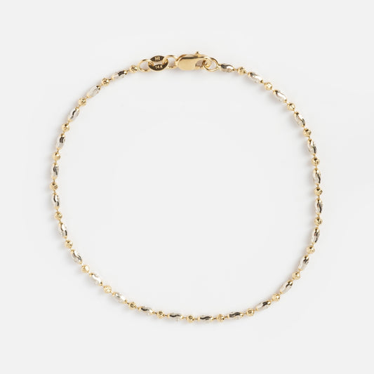 The Bubble Two Tone Bracelet is a delicate gold chain with small bead accents and a lobster clasp, displayed in a circular shape on a plain white background.