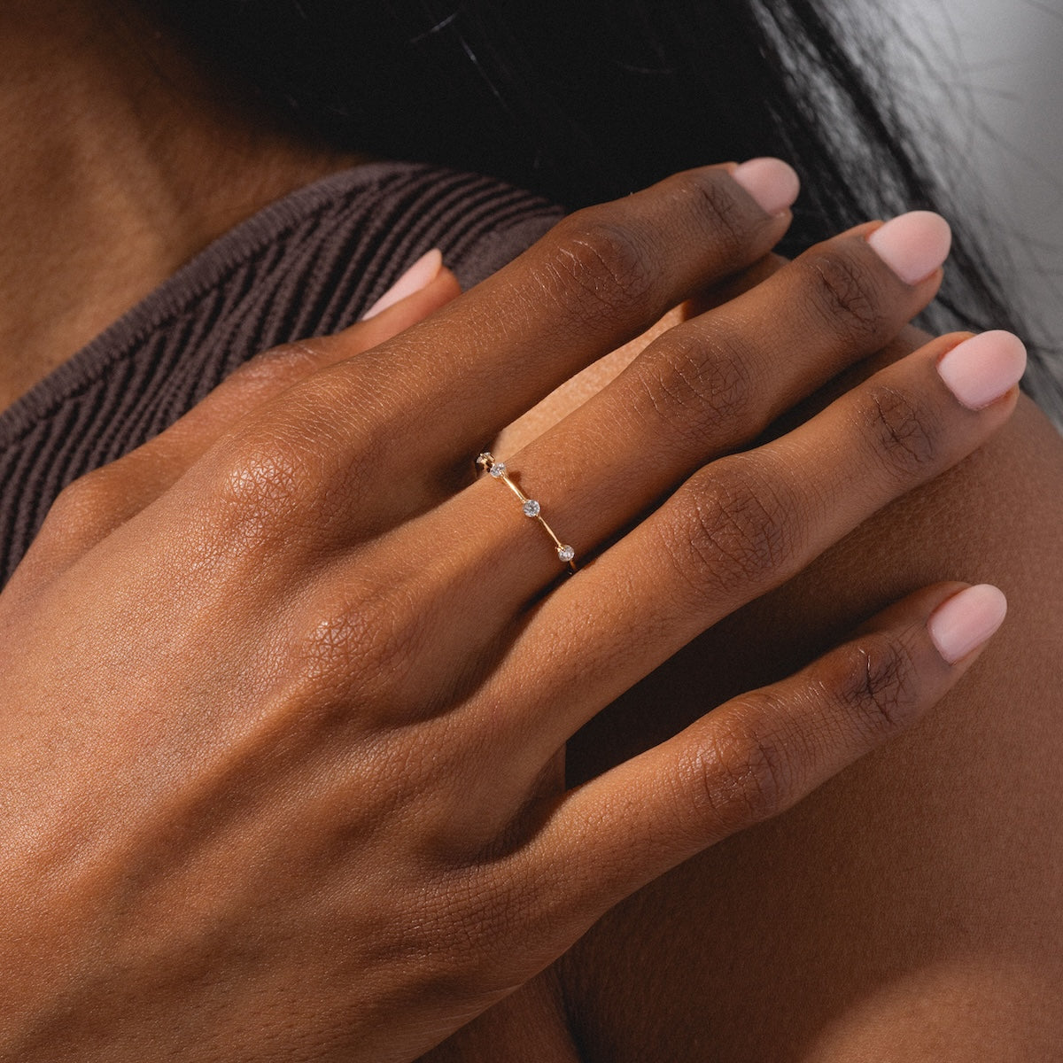 A close-up of a hand with pale pink nail polish wearing the Munro Ring, a delicate gold band adorned with small, spaced diamonds, resting on a textured brown garment.