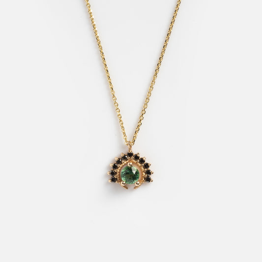 The Tosia Black Diamond Necklace features a gold pendant with a central green gemstone, encircled by small black stones in a semi-circle pattern, set against a plain white background.