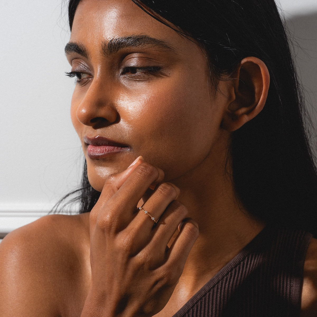 A woman with long dark hair and glowing skin gazes to the side, chin resting on her hand adorned with the Munro Ring. She wears a brown one-shoulder top as sunlight casts soft shadows across her face.