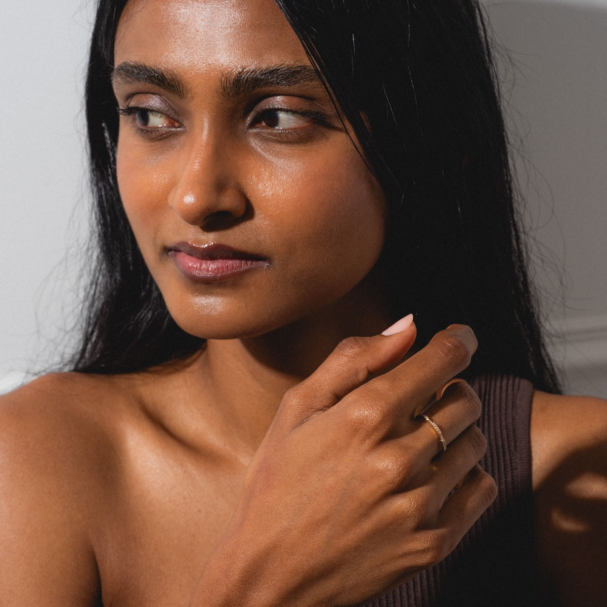 A woman with long dark hair and radiant skin looks to the side, holding her hand by her chin to showcase the simple gold Roxburgh Ring. Sunlight softly highlights her face and hand as she wears a sleeveless brown top.