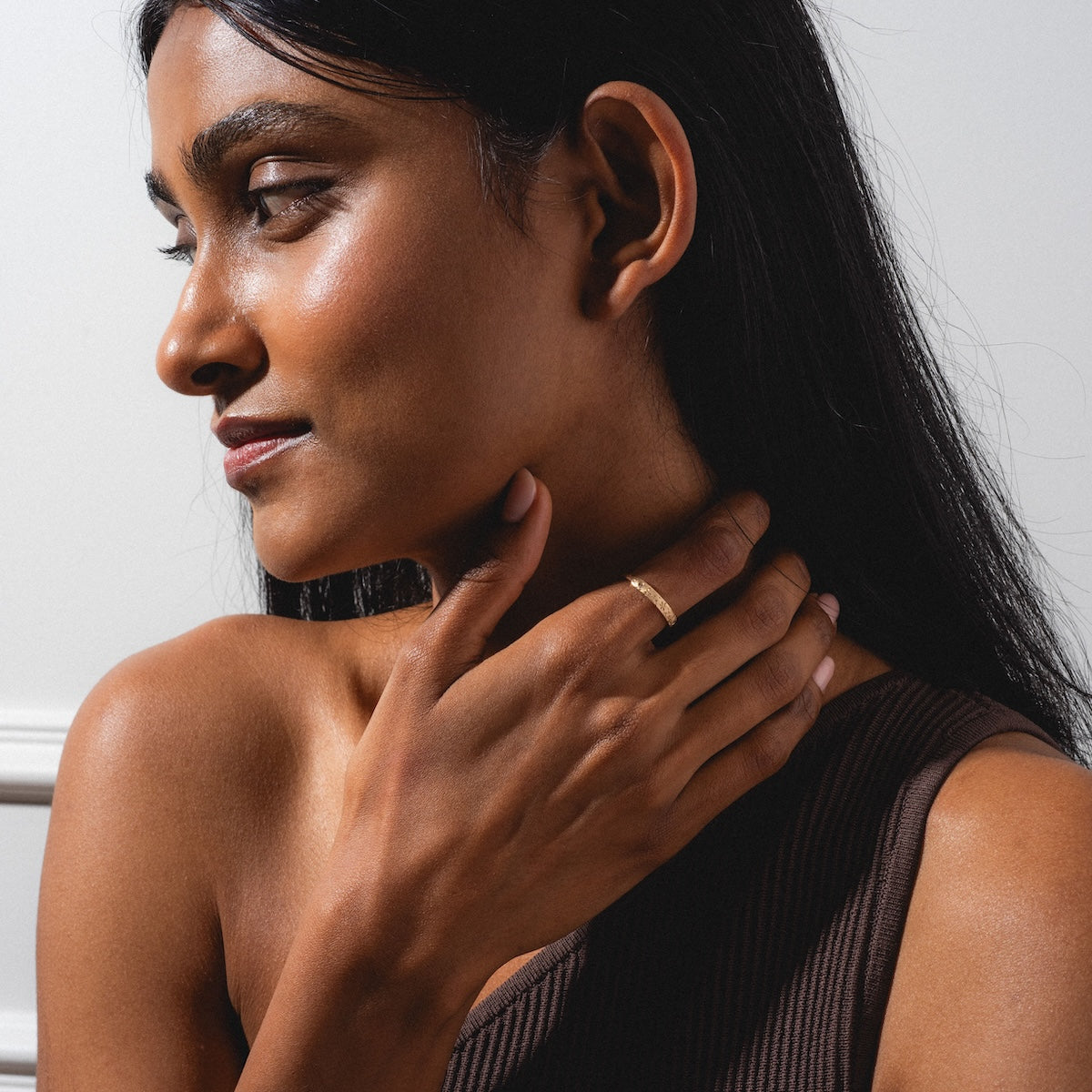 A woman with long dark hair gazes sideways, touching her neck with her left hand. She wears a sleeveless brown top and the Dukes Champagne Diamond Ring, while gentle light highlights her smooth skin.