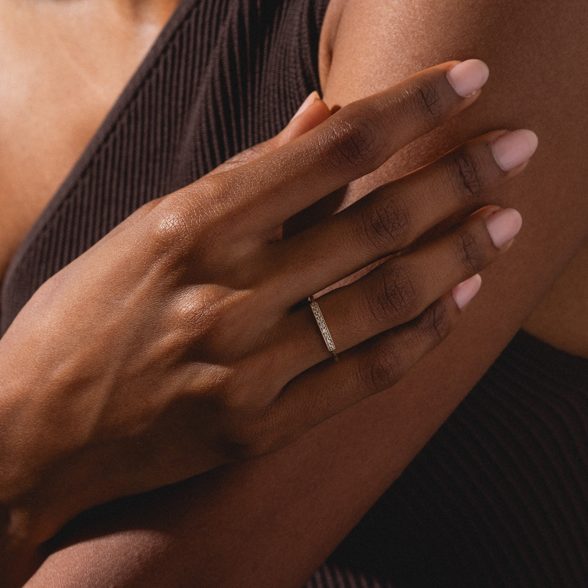 A close-up of a hand with natural nails wearing the thin, gold Clyde Ring, resting gently on the person's opposite arm dressed in ribbed, dark brown fabric.