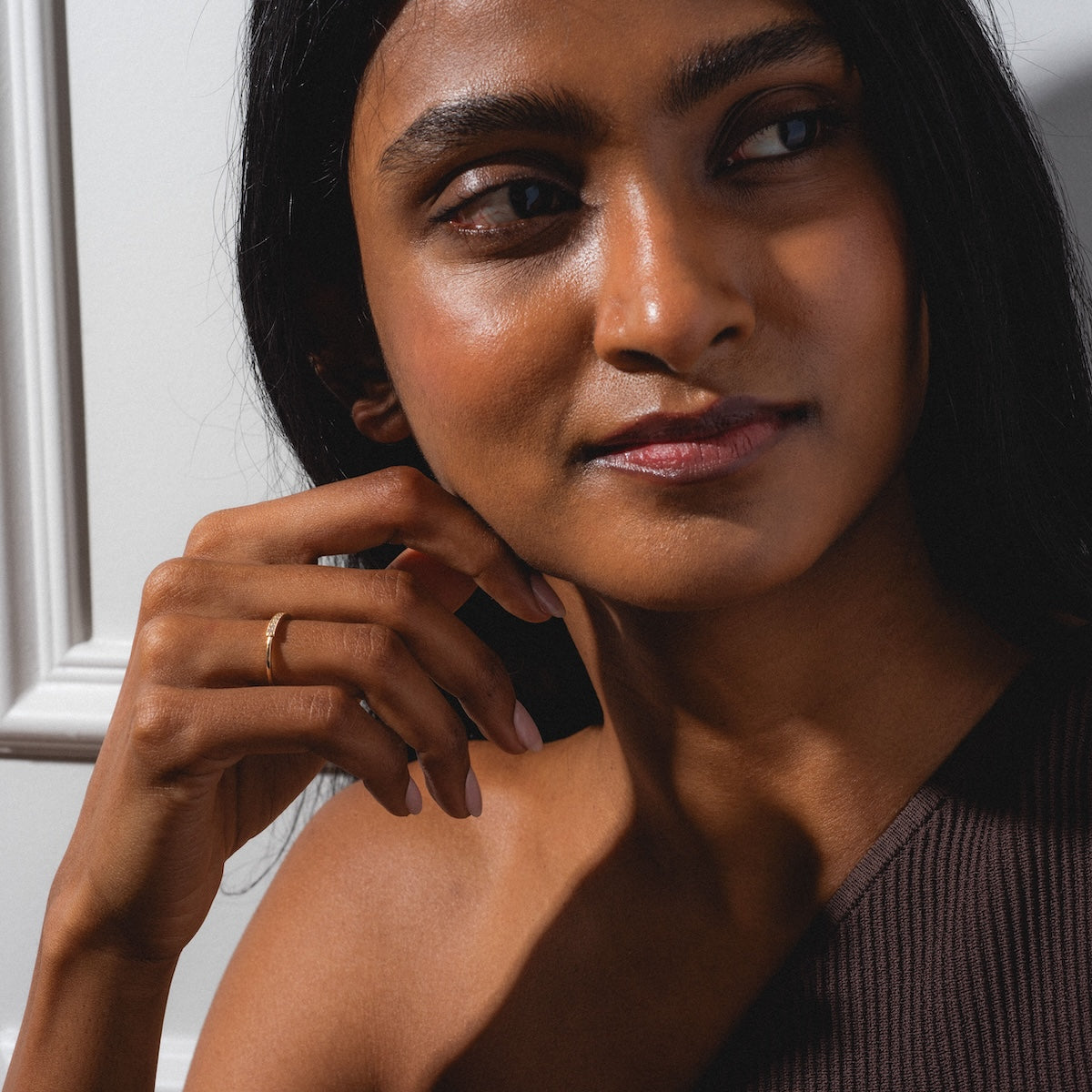 A woman with long dark hair and smooth brown skin gazes to the side, her hand near her chin. She wears a one-shoulder brown top and the Clyde Ring in gold, with sunlight casting gentle shadows on her face.