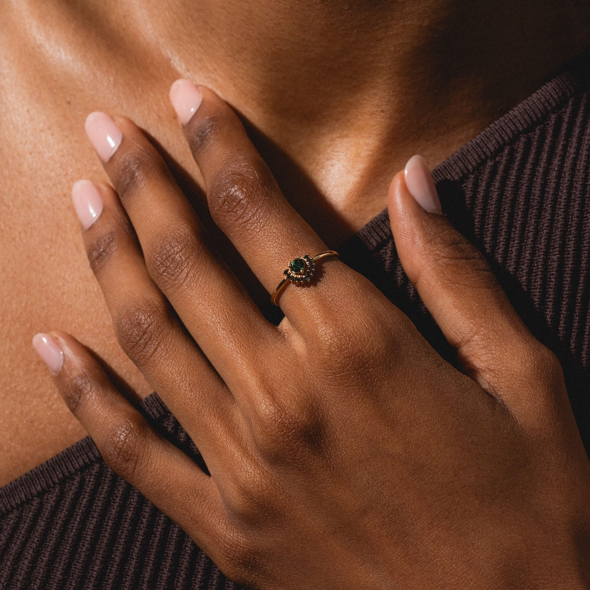 A close-up of a hand with pale pink nails gently resting on someone’s chest, showcasing the Tosia Tourmaline Ring. The person is wearing a dark ribbed top.