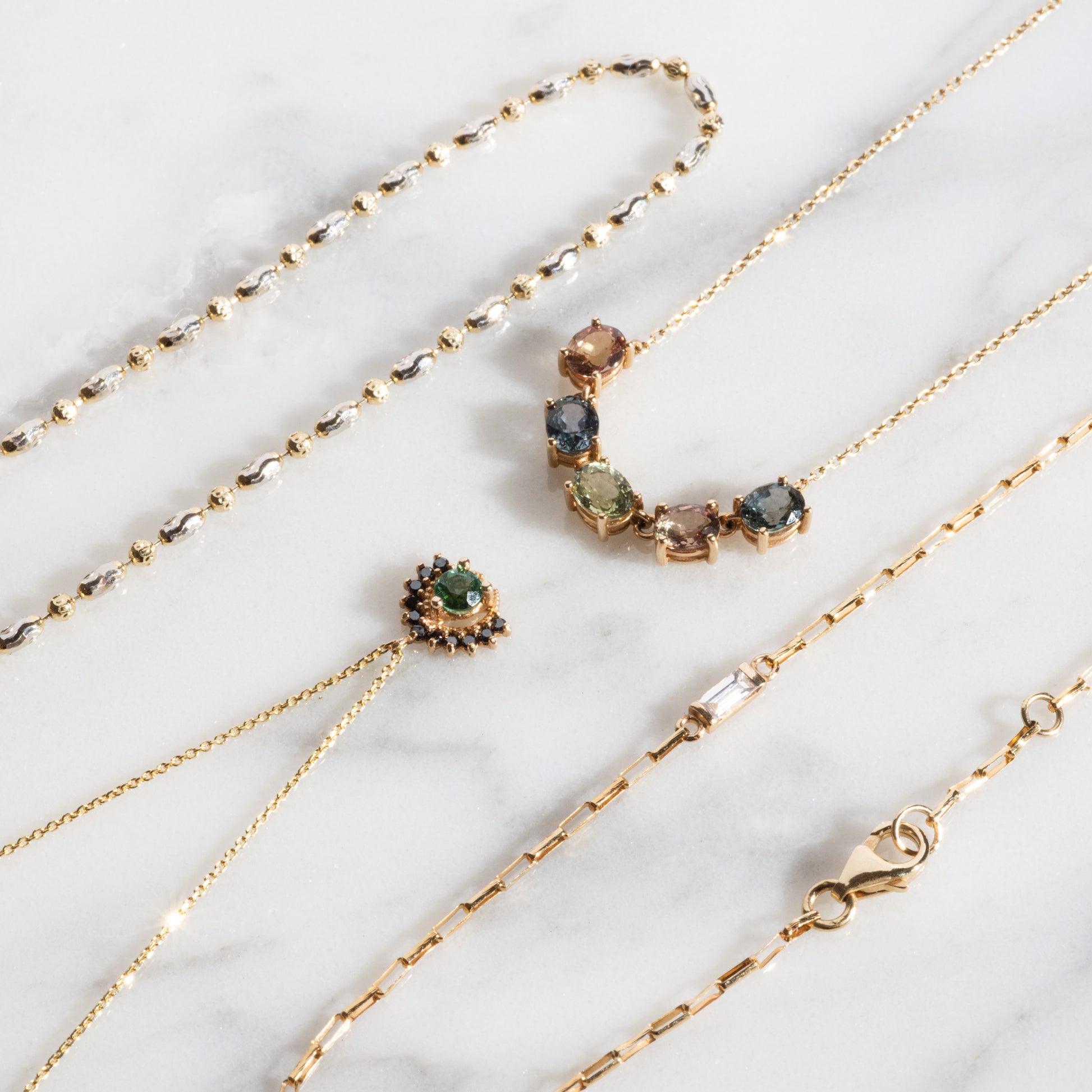 Four Royal Mile Necklaces with gold chains and assorted gemstone and bead designs are displayed on a white marble surface, highlighting their intricate links and vibrant stones.