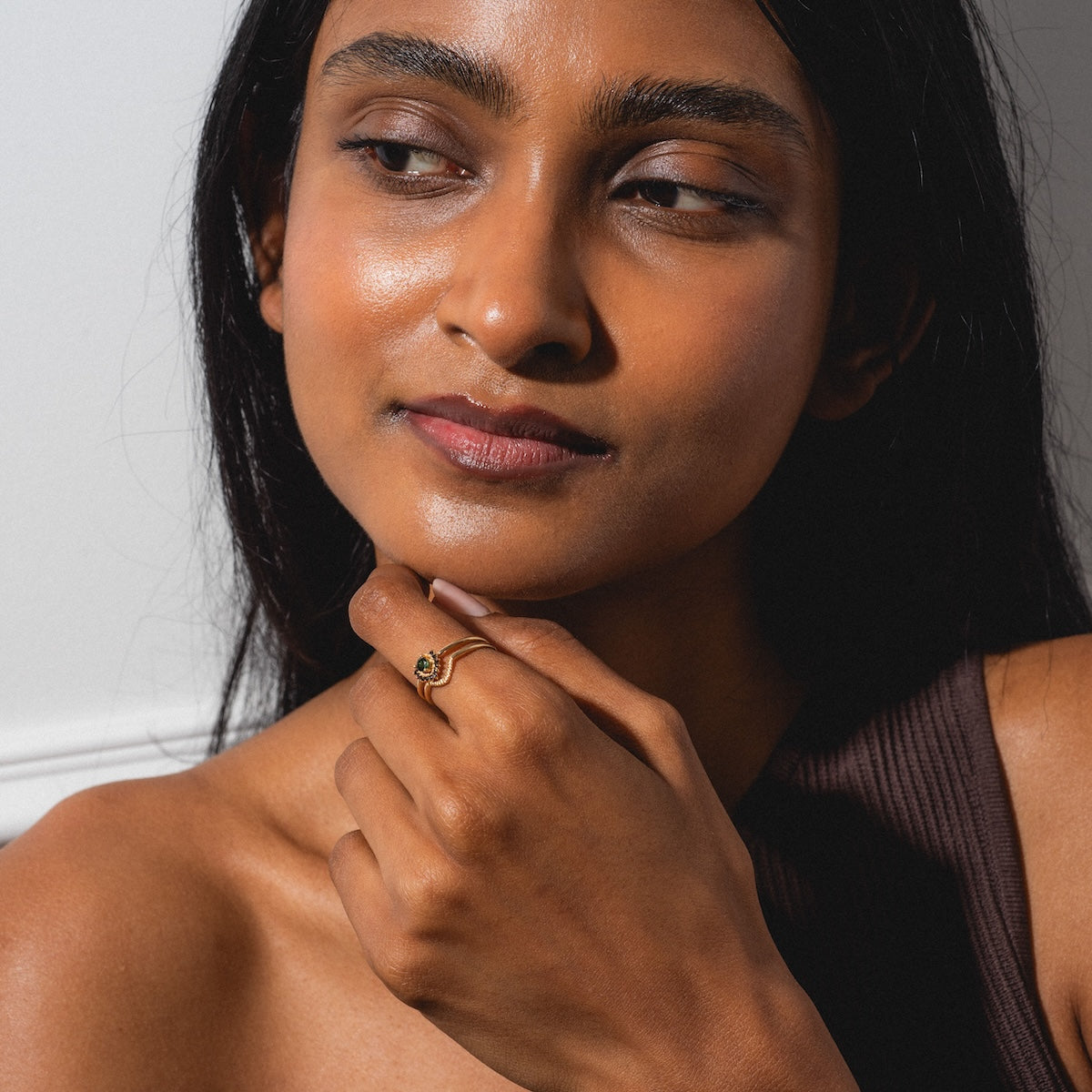 A woman with long dark hair wears a sleeveless brown top and gazes thoughtfully to the side, her hand touching her chin and showcasing the Cairn Ring.