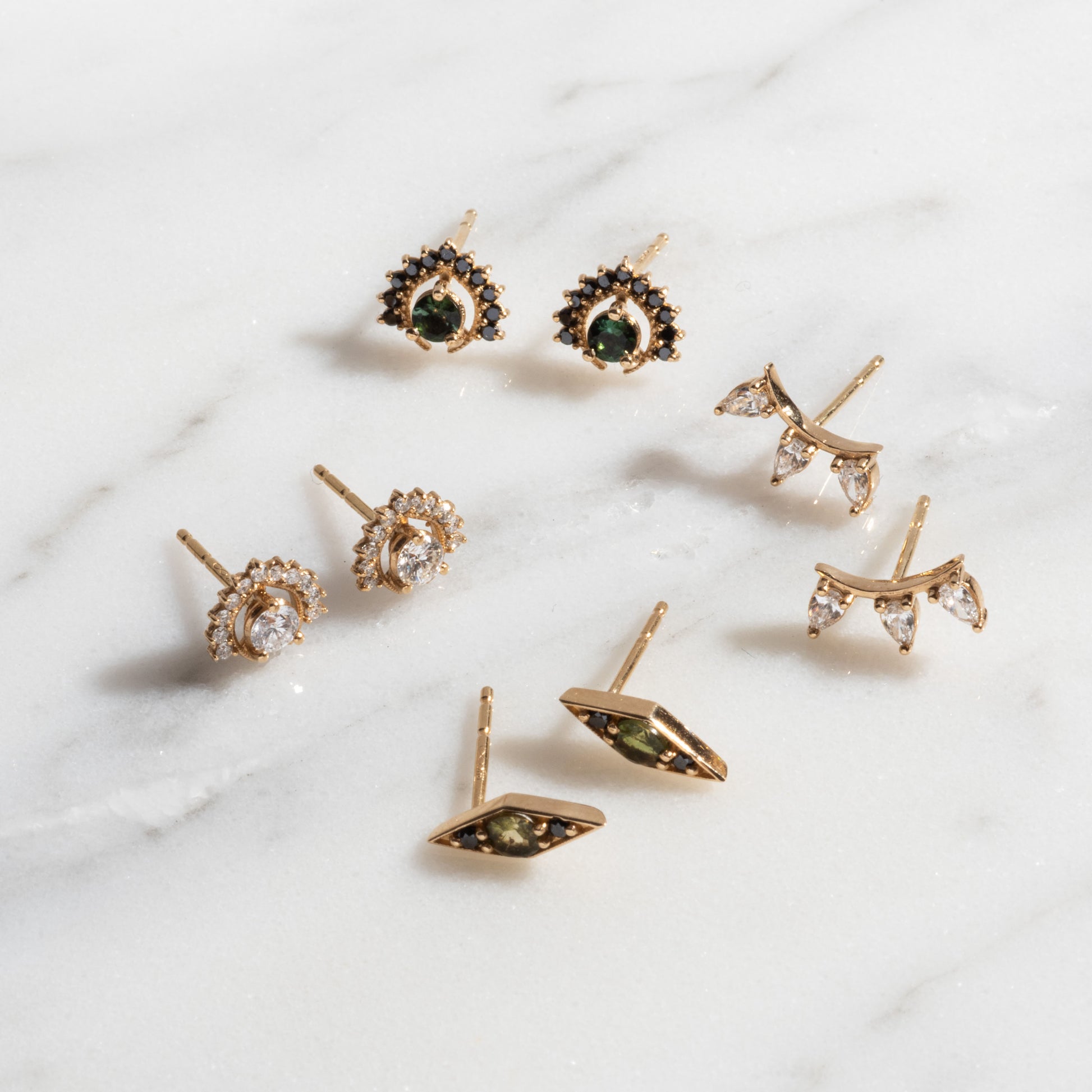 Four pairs of Tosia Diamond Studs in gold, featuring green, black, and clear gem designs, are displayed on a white marble surface.