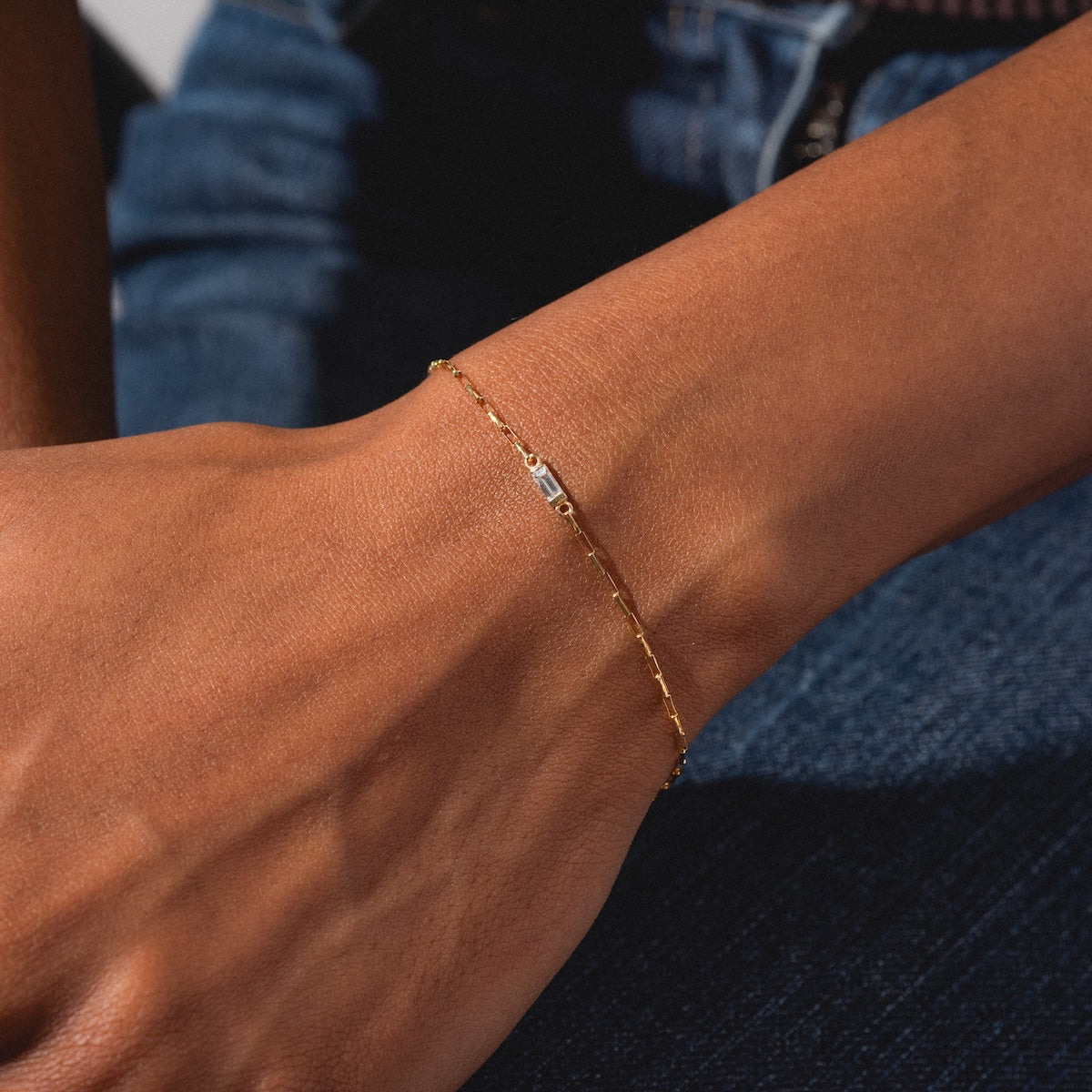 A close-up of a wrist wearing the Sunblink Diamond Bracelet, featuring a delicate gold band with a small rectangular charm, set against blue denim jeans.