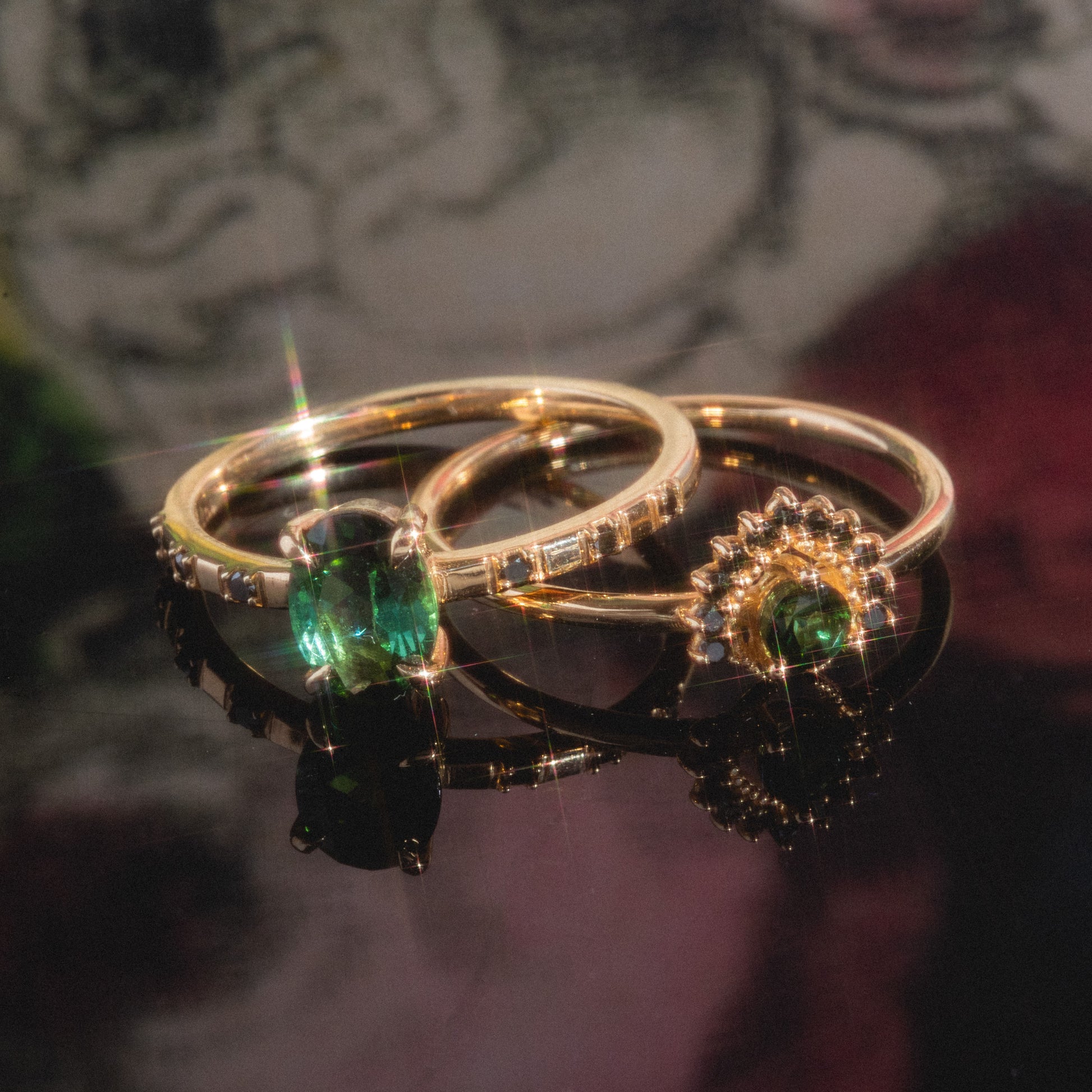 Two Tosia Tourmaline Rings gleam side by side on a reflective surface; one showcases a faceted oval green tourmaline, while the other features a smaller gem in an elegant starburst setting accented with black diamond sparkle.