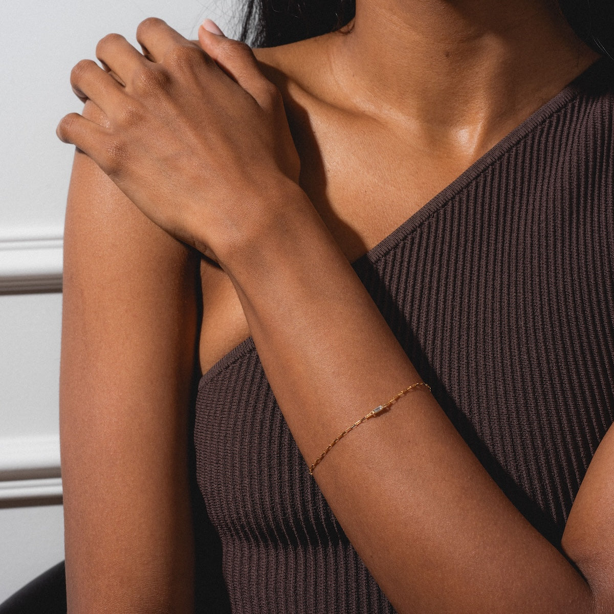 A woman wearing a one-shoulder ribbed brown top poses with her hand on her opposite shoulder, showcasing the Sunblink Diamond Bracelet on her wrist against a light background.