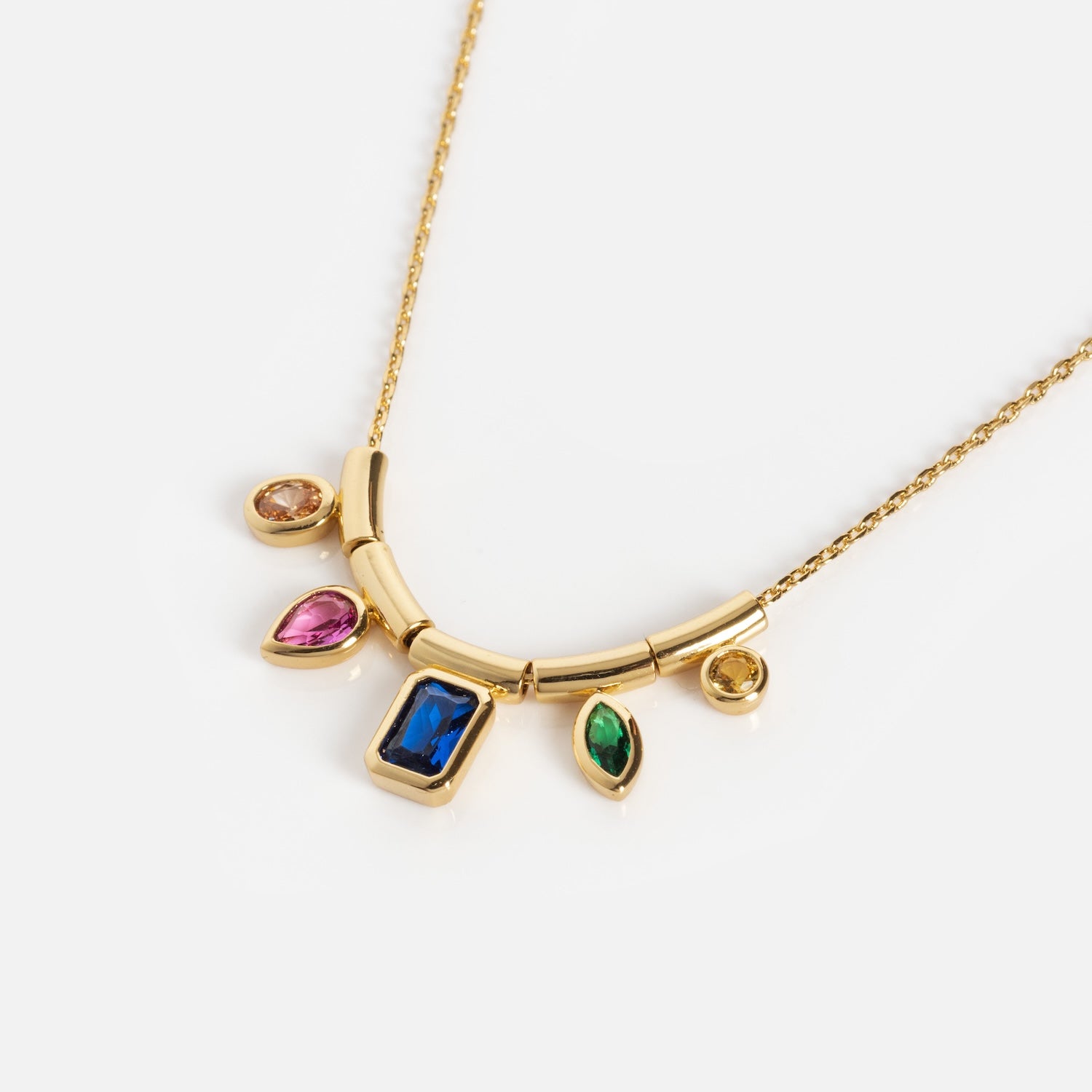 The Technicolor Gem Drop Necklace features a curved gold bar with five multicolored gemstone charms—blue, green, pink, and yellow—dangling from a delicate gold chain, shown on a white background.