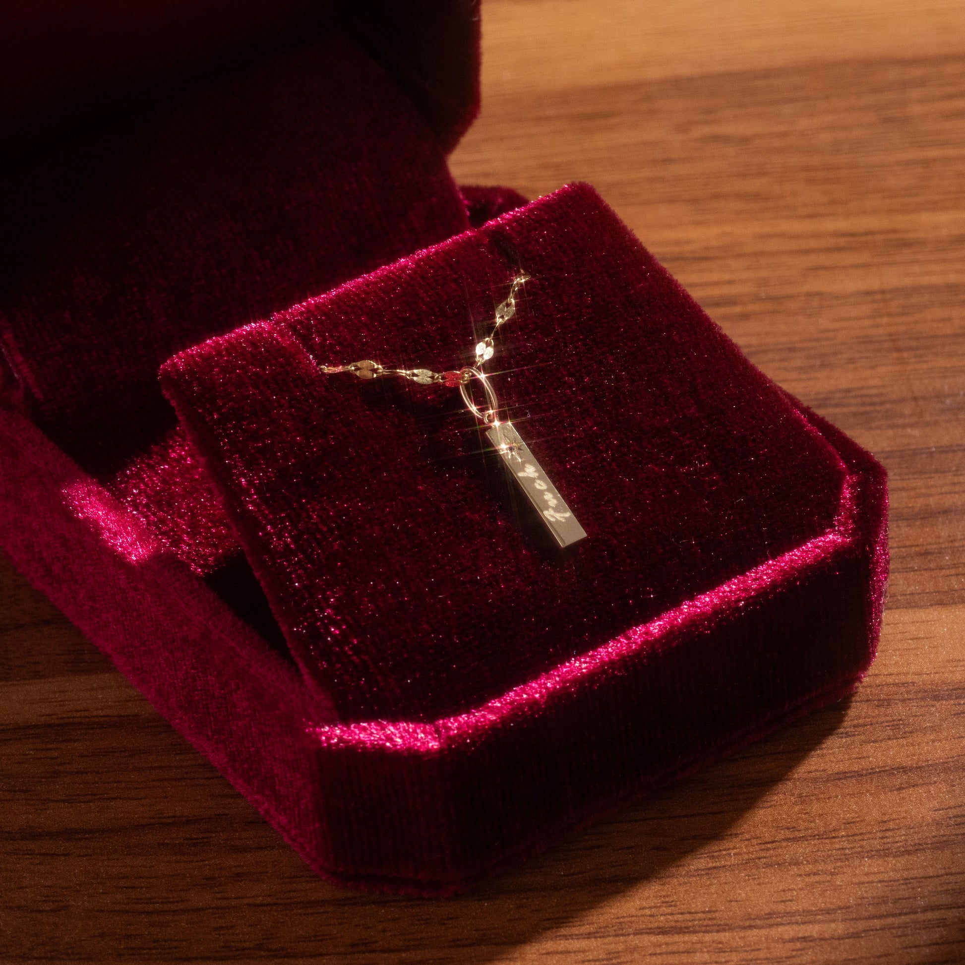 A Solid Gold Tiny Fuck Charm necklace with a rectangular pendant, displayed in a red velvet jewelry box on a wooden surface. The pendant is engraved with the word FUCK.
