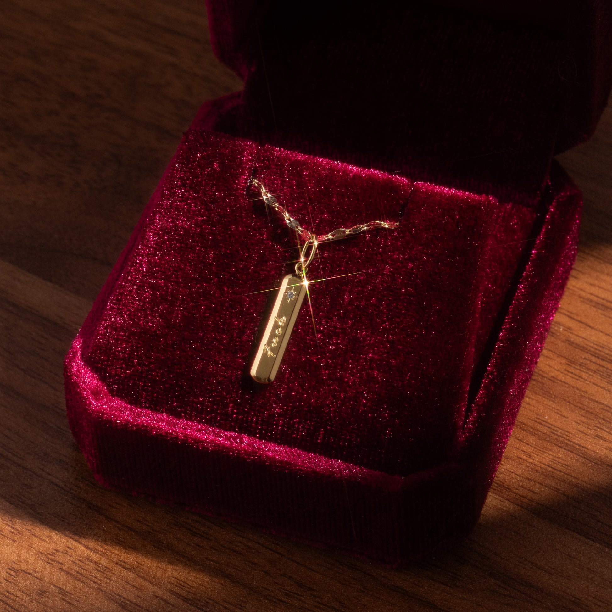 The Tiny Fuck Charm, a gold bar pendant necklace with a delicate chain, sits in an open red velvet jewelry box on a wooden surface, illuminated by soft, warm light.