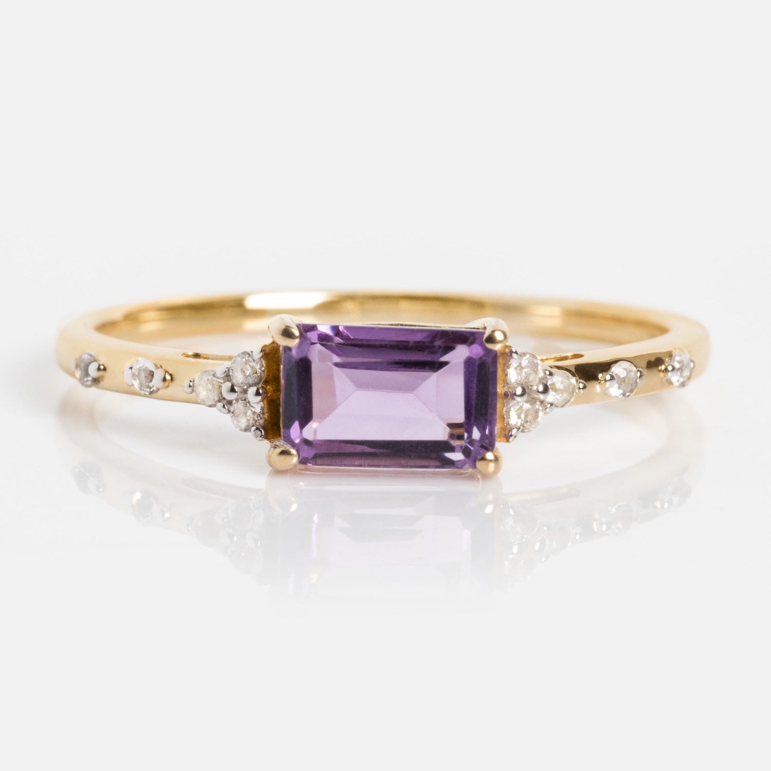 The Solid Gold Glimmering Birthstone Baguette Ring showcases a rectangular purple gemstone at the center, accented by round diamonds along the band, and is displayed on a white background.
