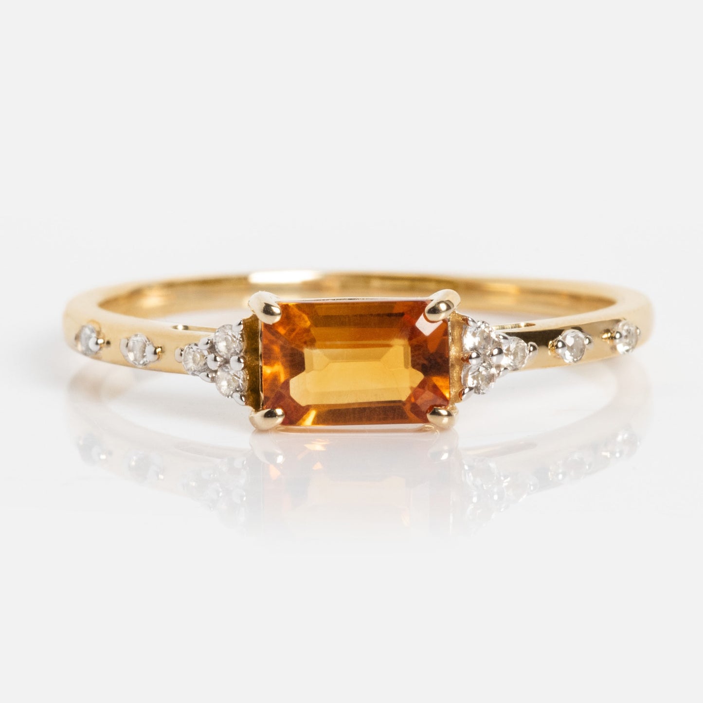 The Solid Gold Glimmering Birthstone Baguette Ring showcases a rectangular orange gemstone at the center with sparkling white topaz accents on each side, set against a plain white background.