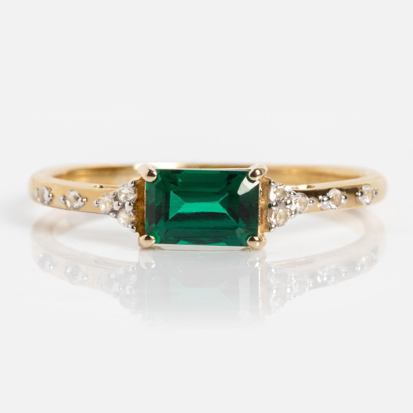 The Solid Gold Glimmering Birthstone Baguette Ring showcases a rectangular emerald-cut green gemstone at the center, accented by round diamonds on each side along the solid gold band.