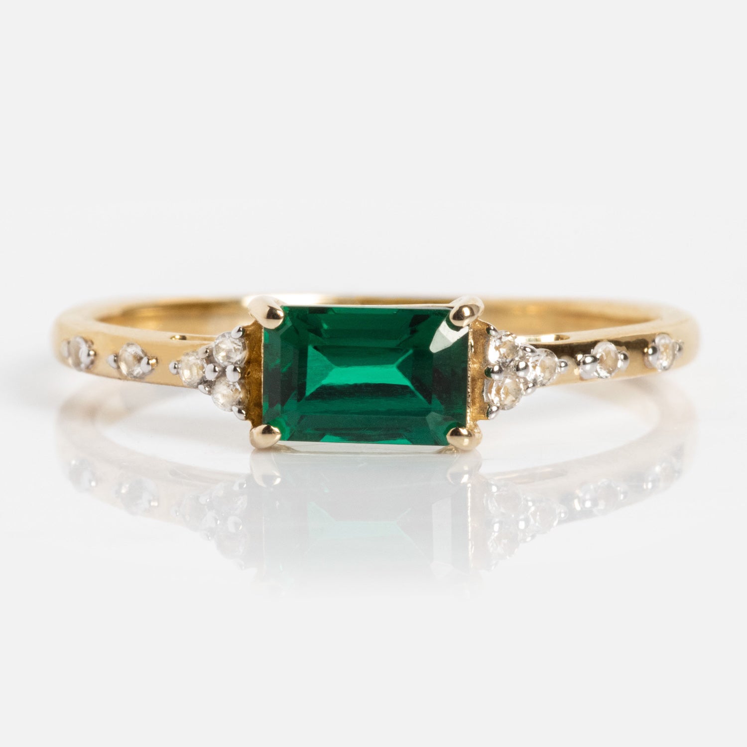 The Solid Gold Glimmering Birthstone Baguette Ring showcases a rectangular emerald-cut green gemstone at the center, accented by round diamonds on each side along the solid gold band.