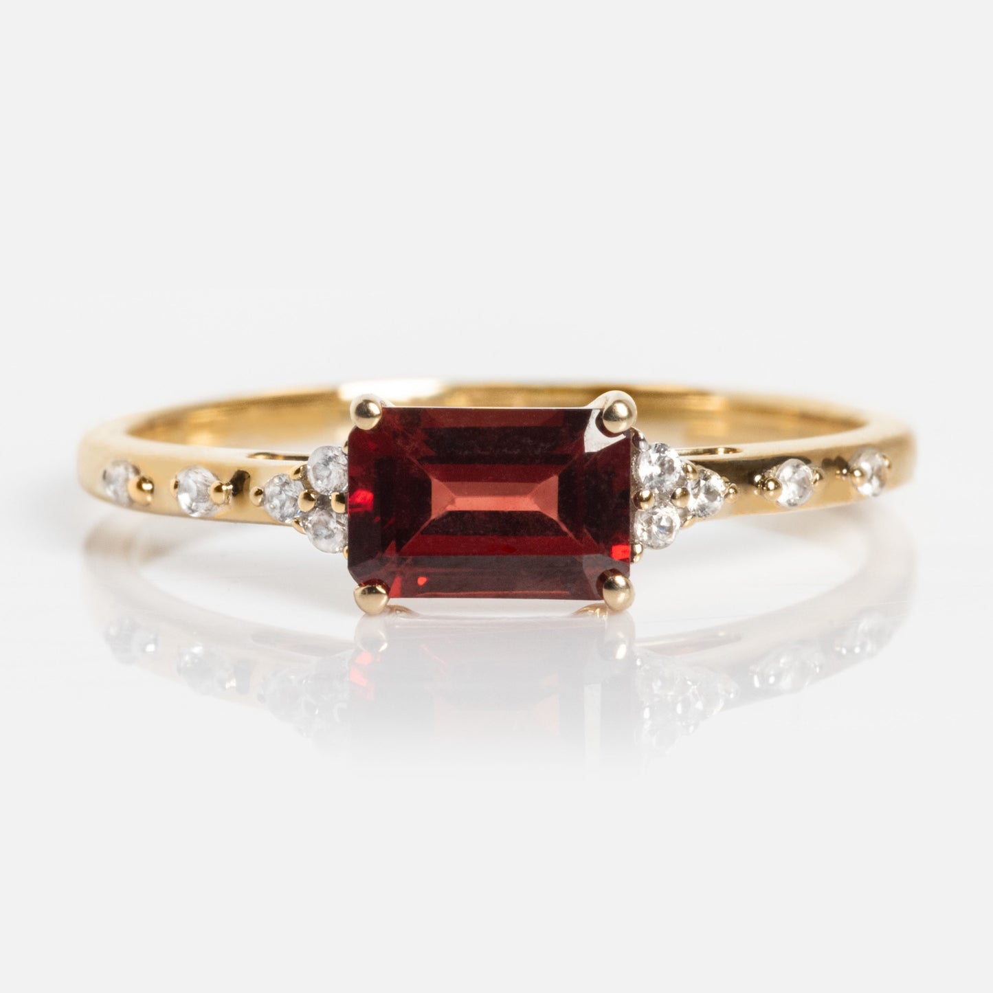 The Solid Gold Glimmering Birthstone Baguette Ring showcases a rectangular red baguette gemstone at the center, accented by small round white stones along the band, all presented on a white background.