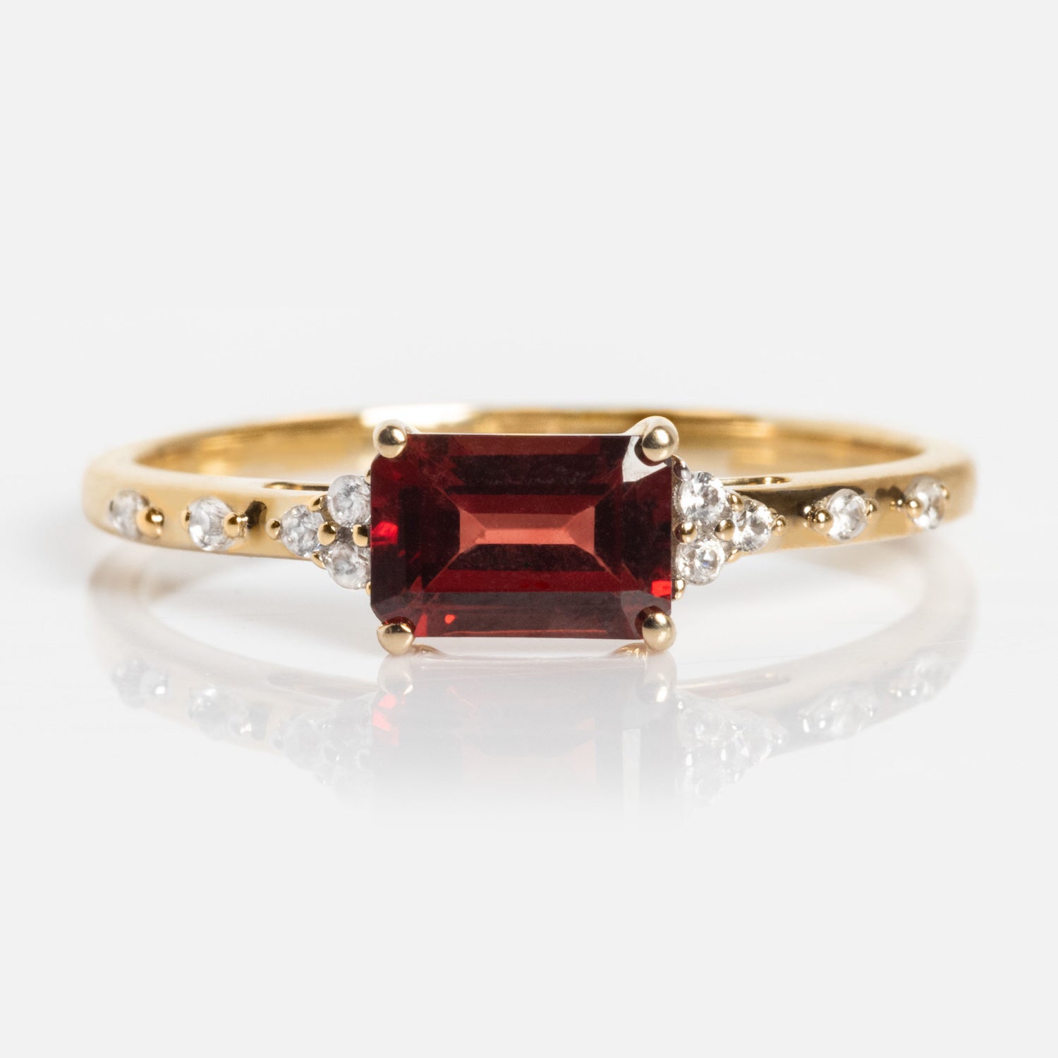 The Solid Gold Glimmering Birthstone Baguette Ring showcases a rectangular red baguette gemstone at the center, accented by small round white stones along the band, all presented on a white background.