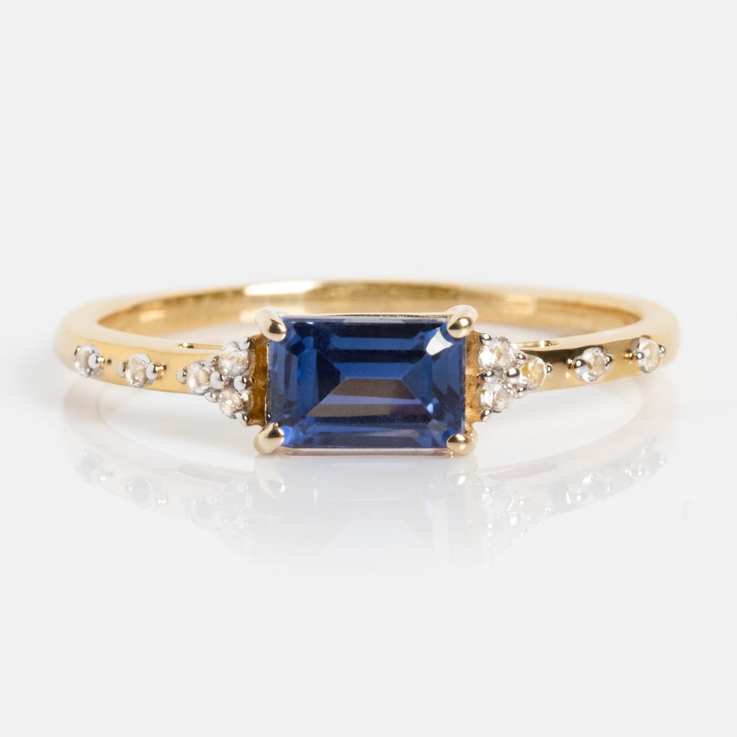 The Solid Gold Glimmering Birthstone Baguette Ring features a rectangular birthstone center, accented by round white topaz on each side and additional white stones along the band.