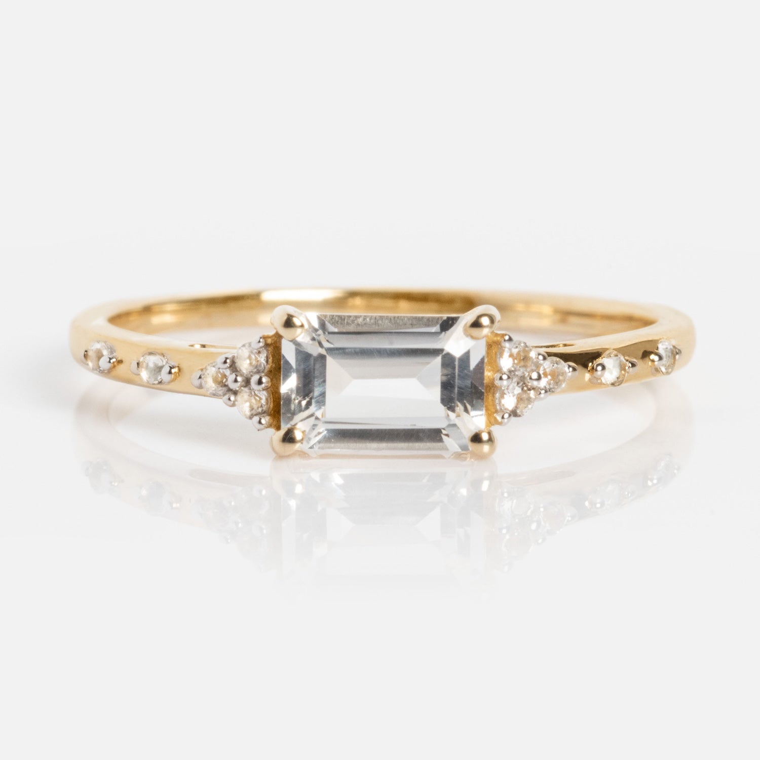 The Solid Gold Glimmering Birthstone Baguette Ring features a rectangular, emerald-cut clear center stone with small round diamonds and white topaz accents set on a slender gold band.