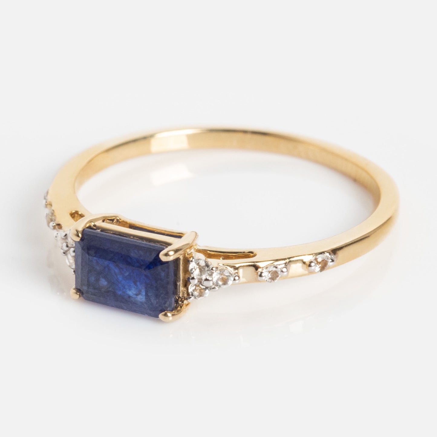 The Solid Gold Glimmering Birthstone Baguette Ring highlights a rectangular blue stone at the center with white topaz accents on both sides, showcased against a plain white background.