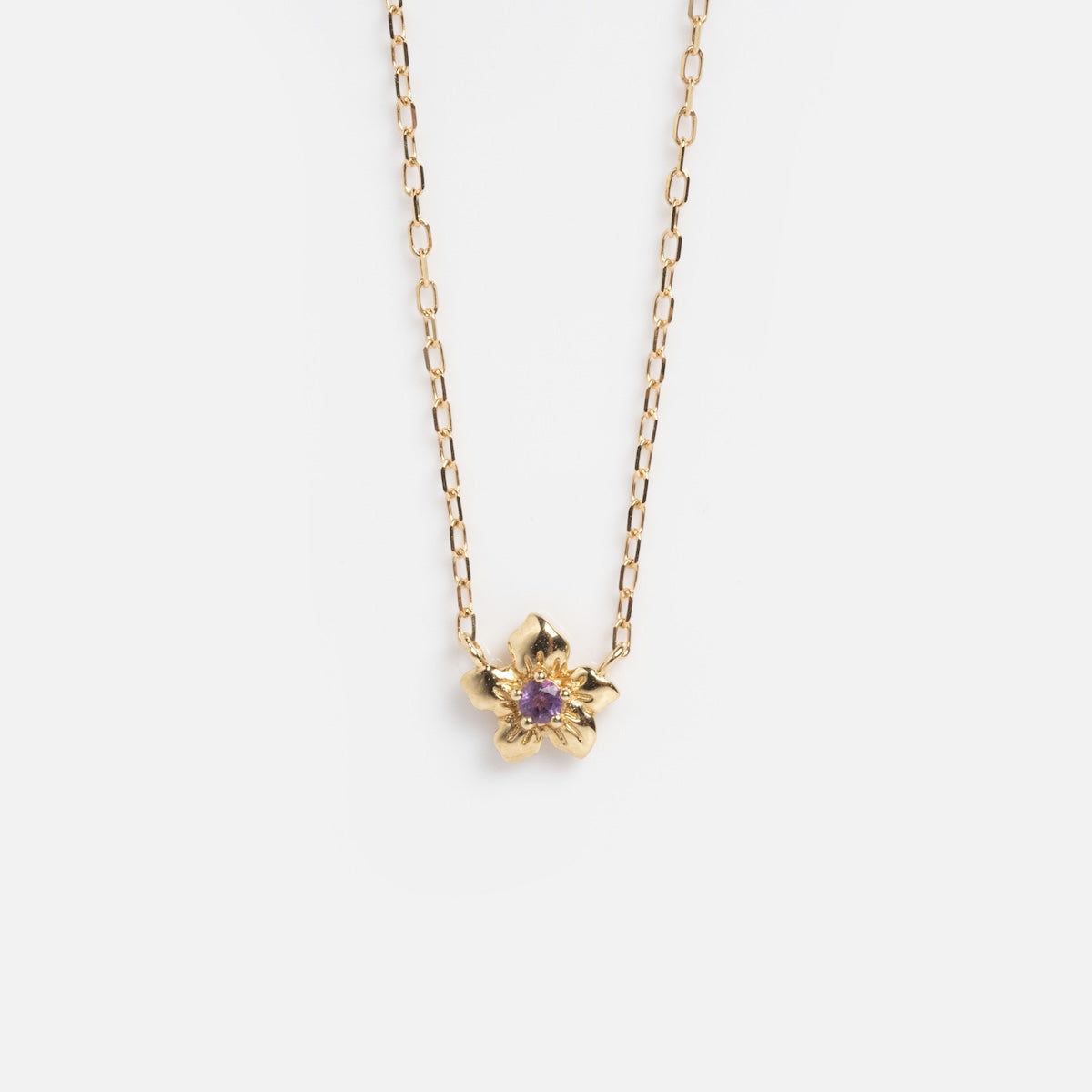 The Solid Gold Birth Flower Bloom Necklace features a delicate flower-shaped pendant with a central purple gemstone, set on a thin gold chain and displayed against a white background.