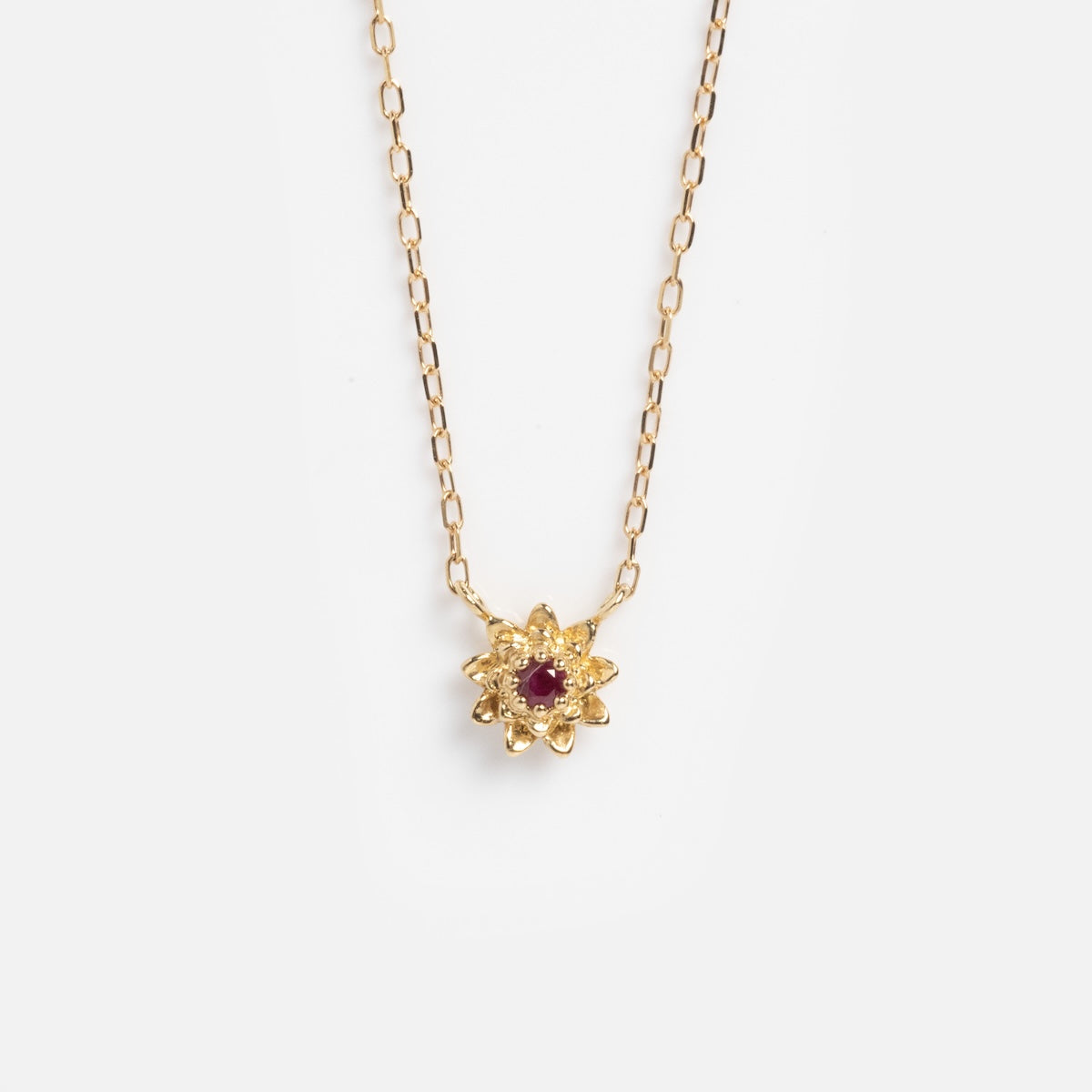 The Solid Gold Birth Flower Bloom Necklace features a delicate gold chain and a small, flower-shaped pendant with a central red gemstone, shown on a plain white background.