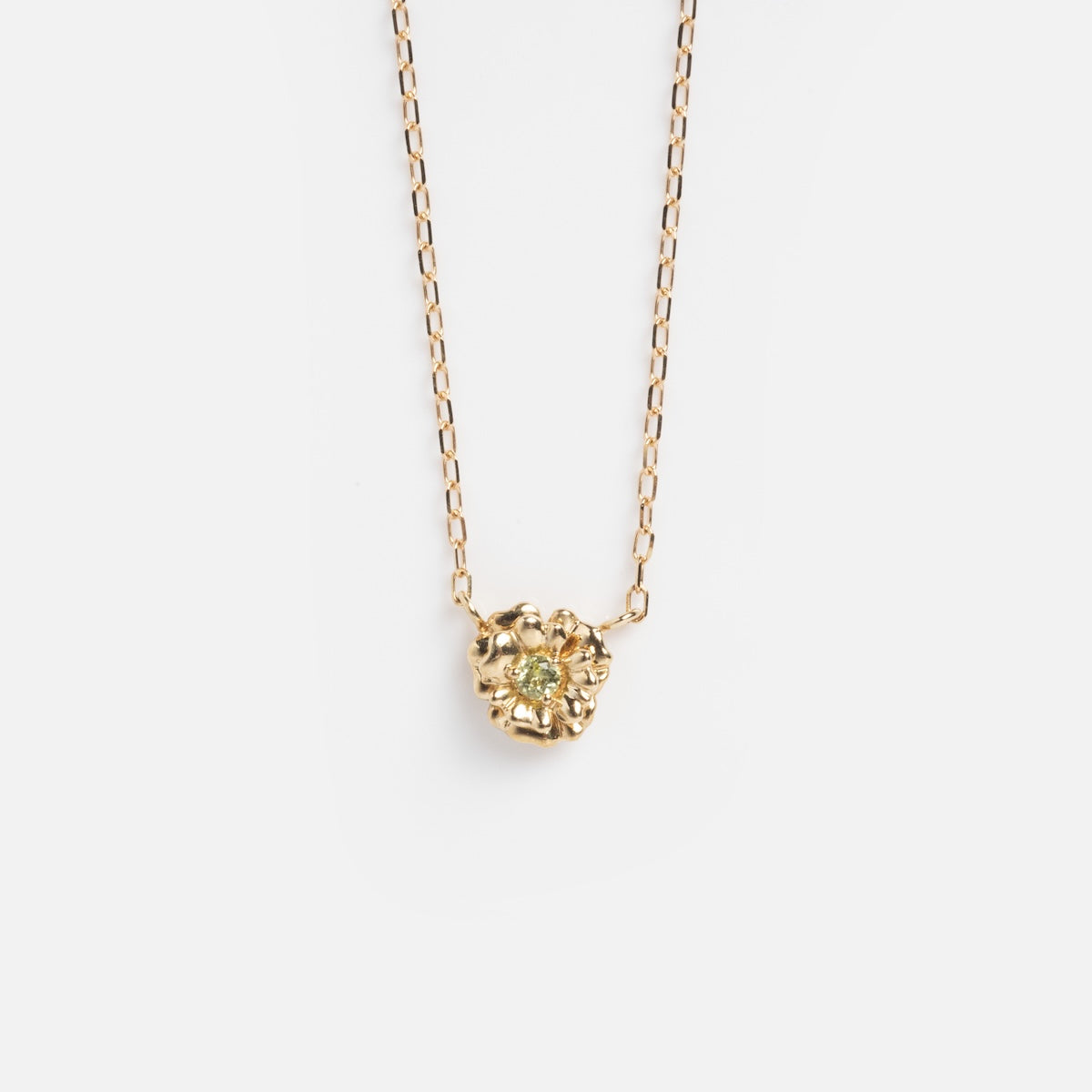 The Solid Gold Birth Flower Bloom Necklace features a delicate gold pendant with a textured, organic shape and a tiny green gemstone at its center, displayed on a simple gold chain against a white background.