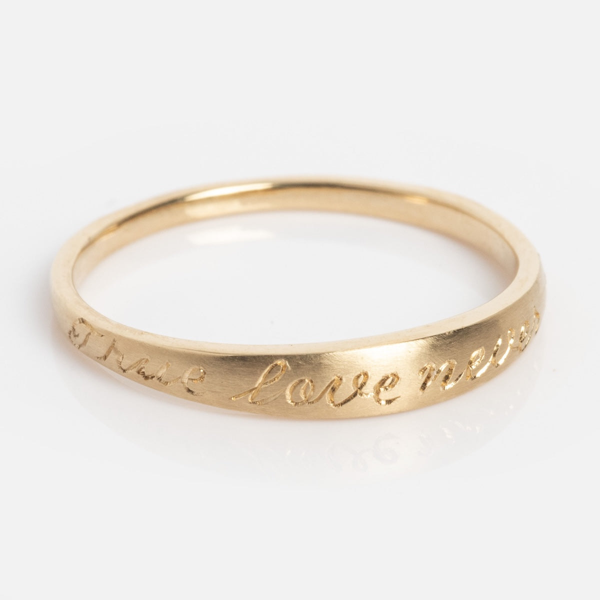 The True Love Never Dies Band is a thin gold ring engraved with “True love never” on its outer surface, shown against a plain white background.
