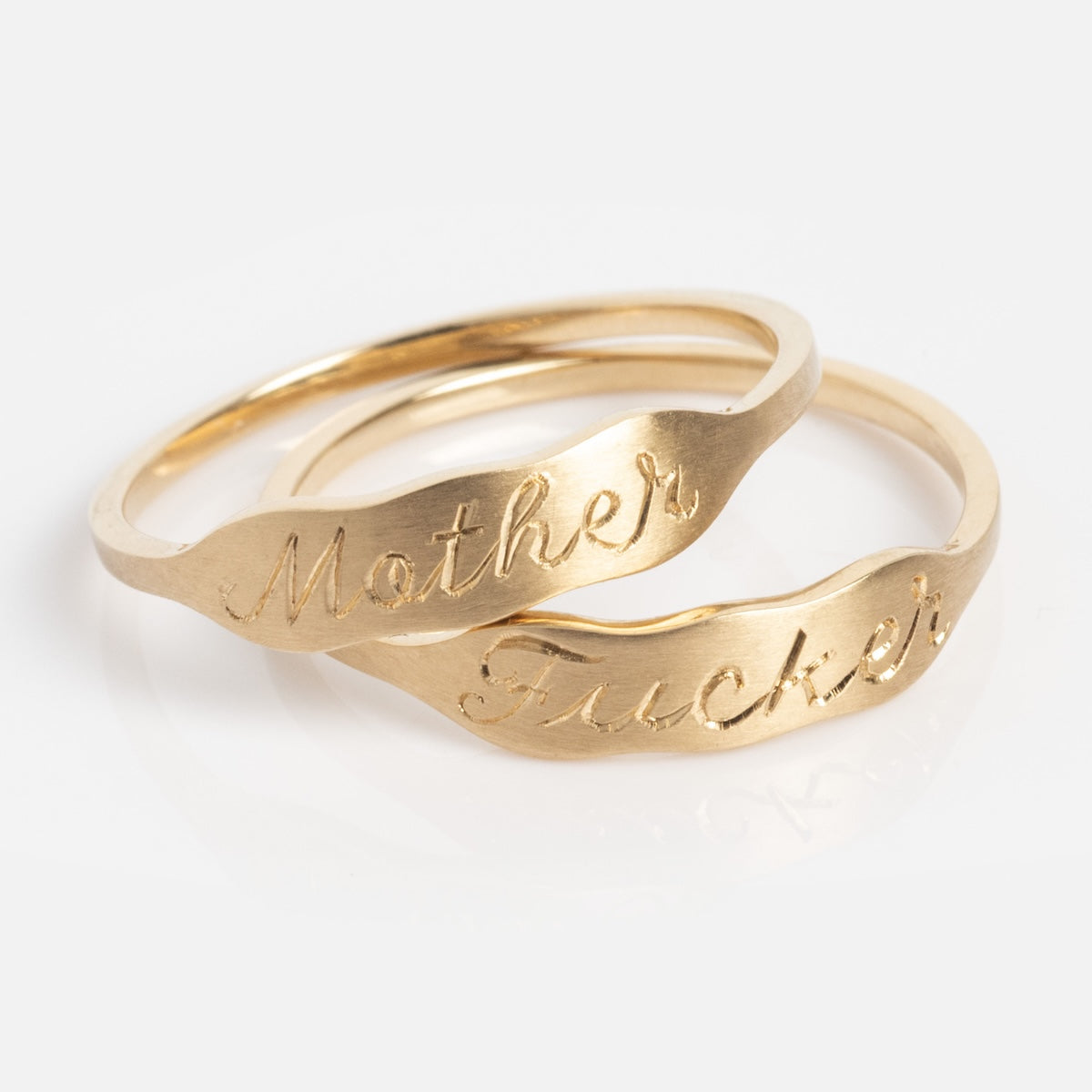 Two 14k Ripples Customizable ID Bands with cursive engravings—one reads "Mother," the other "Fucker." The gold rings overlap on a plain white background.