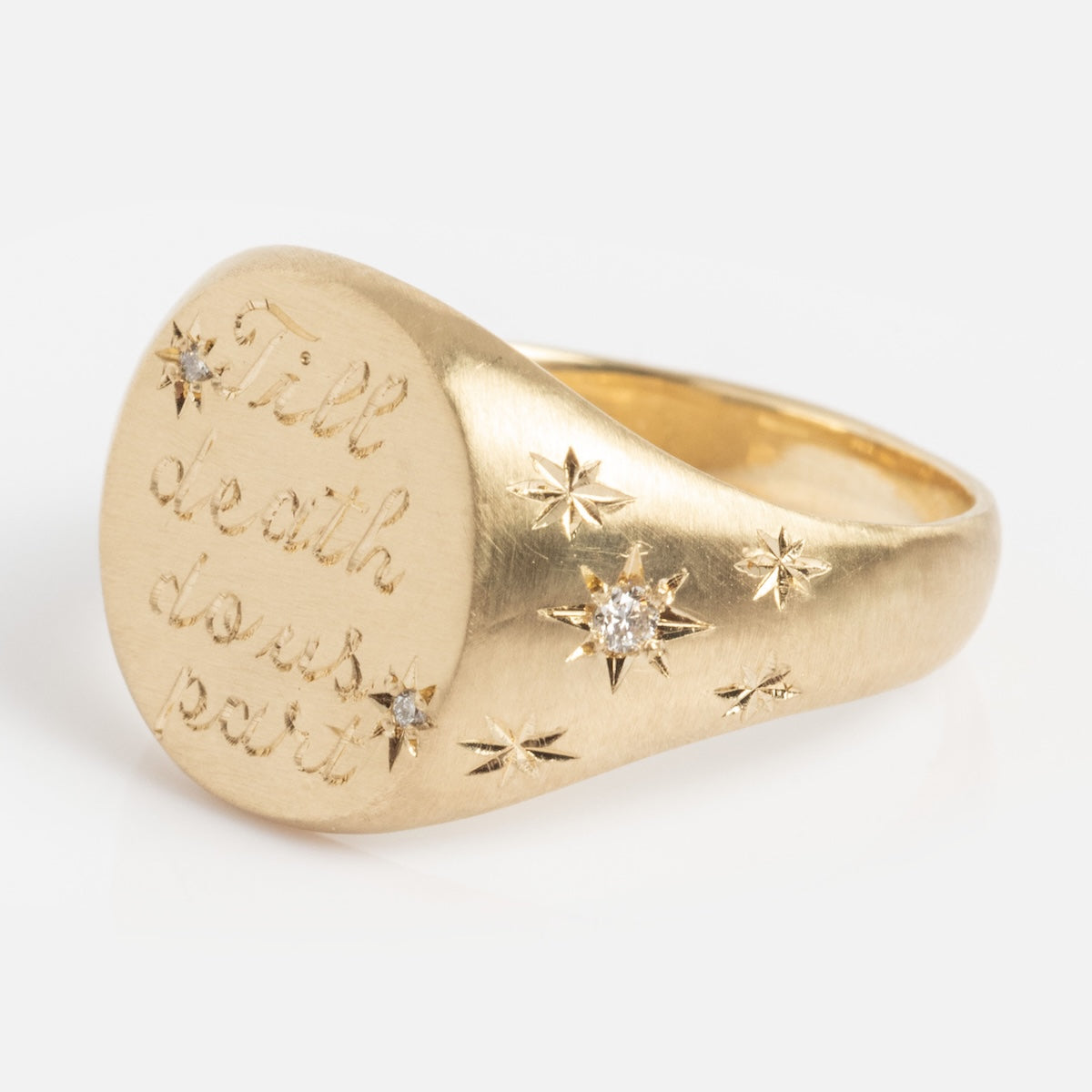 The Till Death Do Us Part Signet with Diamonds is a gold ring engraved with "Till death do us part," featuring star-shaped accents and set with several small diamonds.