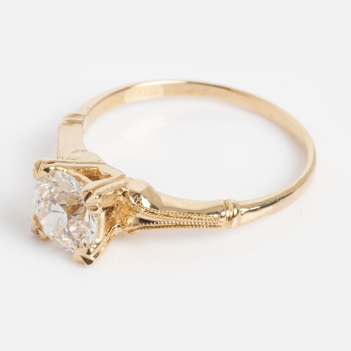 The Elara Ring features a gold band with intricate detailing and a round, faceted diamond in a classic claw setting, shown against a white background.