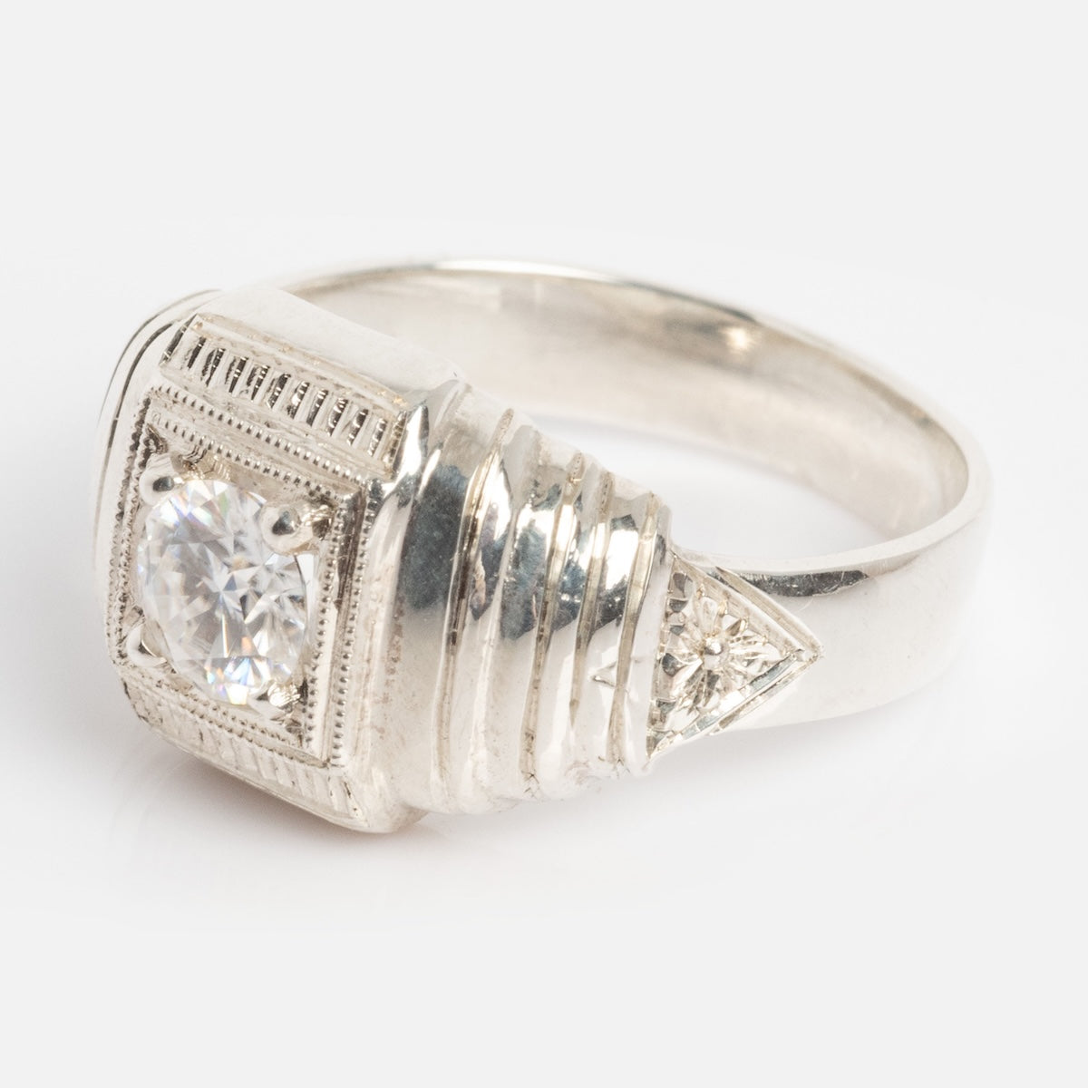 The Sacred Ring is a silver ring with a square, detailed setting, featuring a central round clear gemstone and geometric engravings along the band.