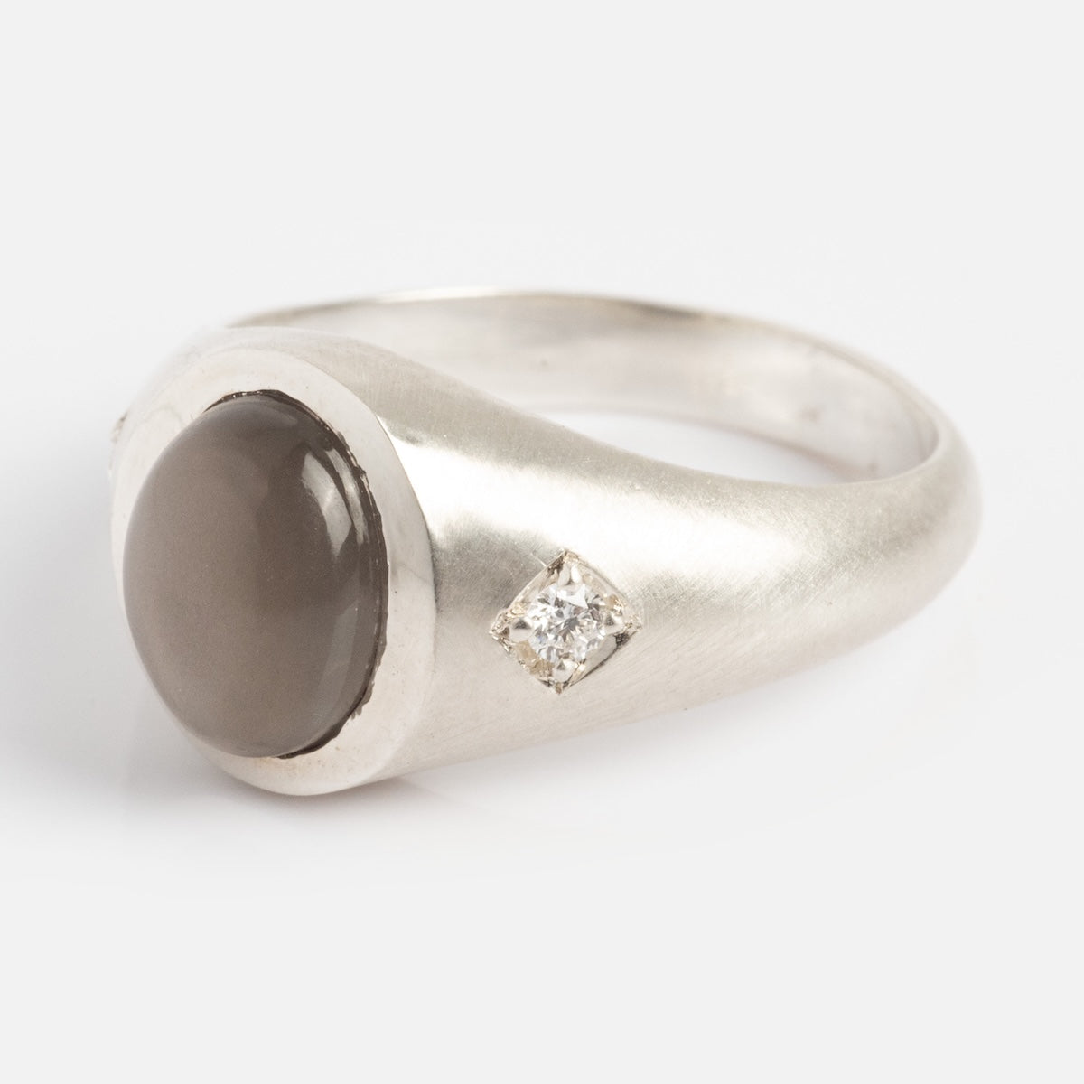The Vailance Ring is a silver band with a round, opaque gray gemstone in a bezel setting, accented by a small, square-set clear diamond beside it.